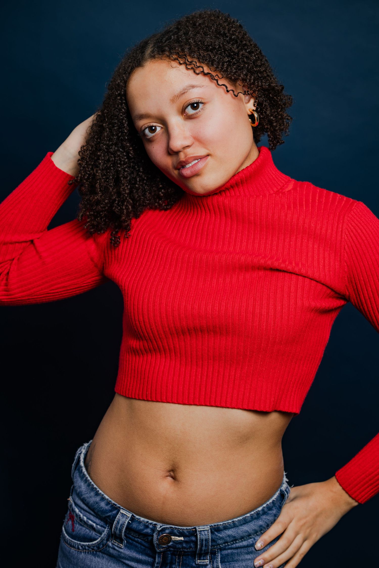 Young woman in a red crop top and jeans poses against a dark blue background.