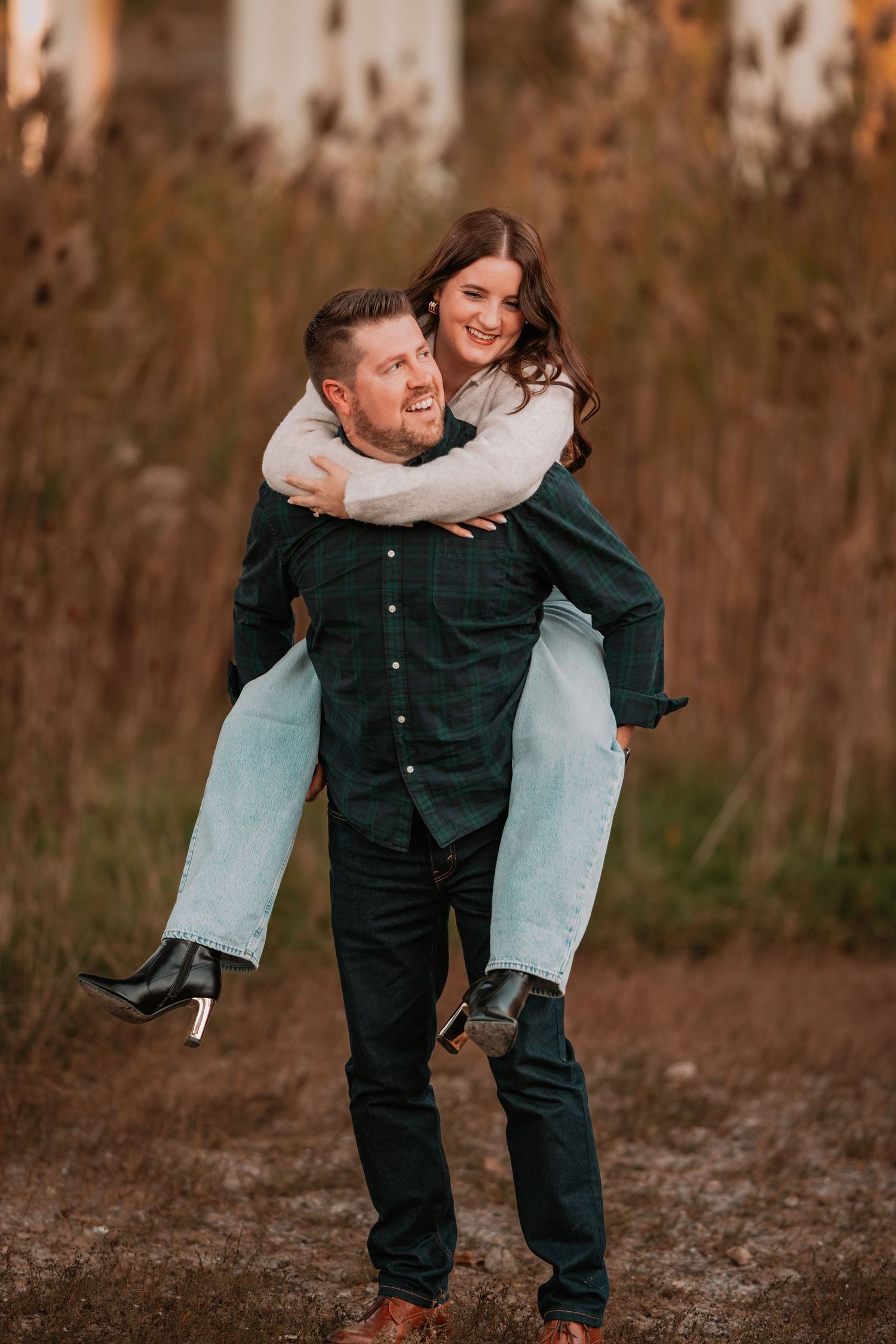 A laughing couple enjoys a piggyback ride in an autumn field, both wearing casual fall outfits.