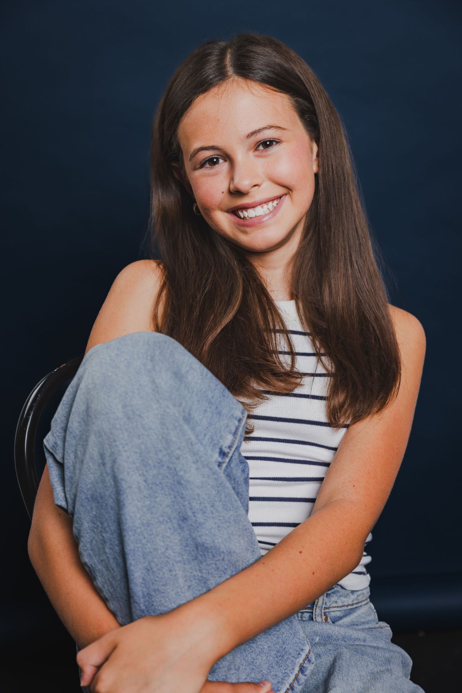 Smiling teenage girl with long brown hair wearing striped top and jeans, sitting casually against dark blue background.