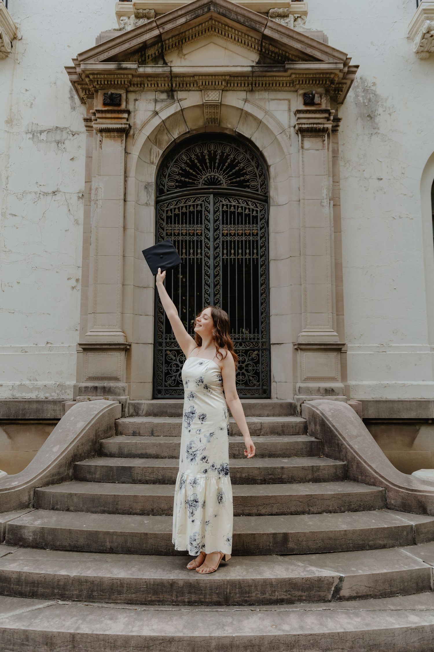 Female graduate in floral dress raising cap triumphantly on steps of ornate building.