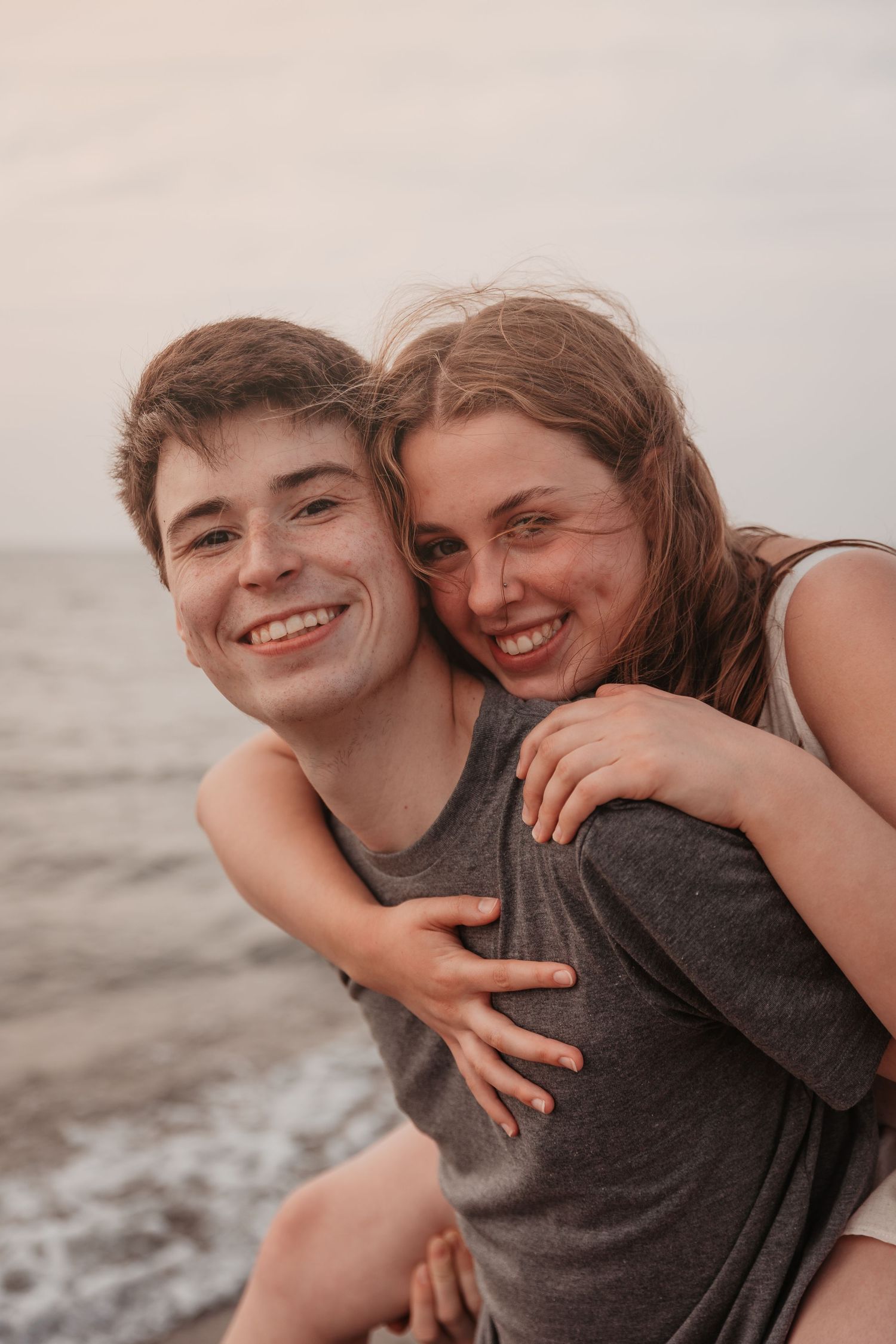 Joyful young couple smiling at beach, woman giving man a piggyback ride near ocean waves.