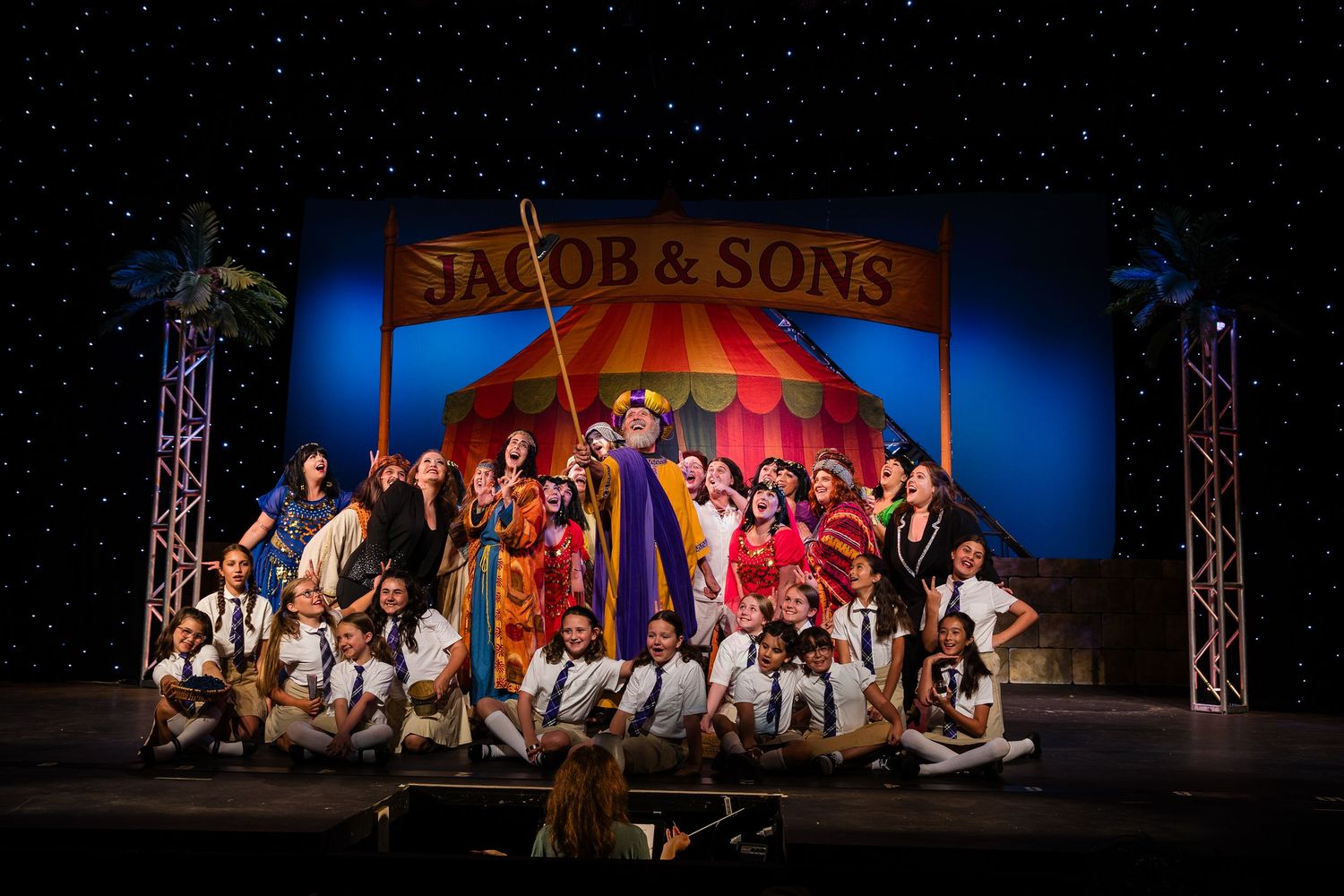 Large cast of Joseph and the Amazing Technicolor Dreamcoat performing on a colorful theatrical stage set.