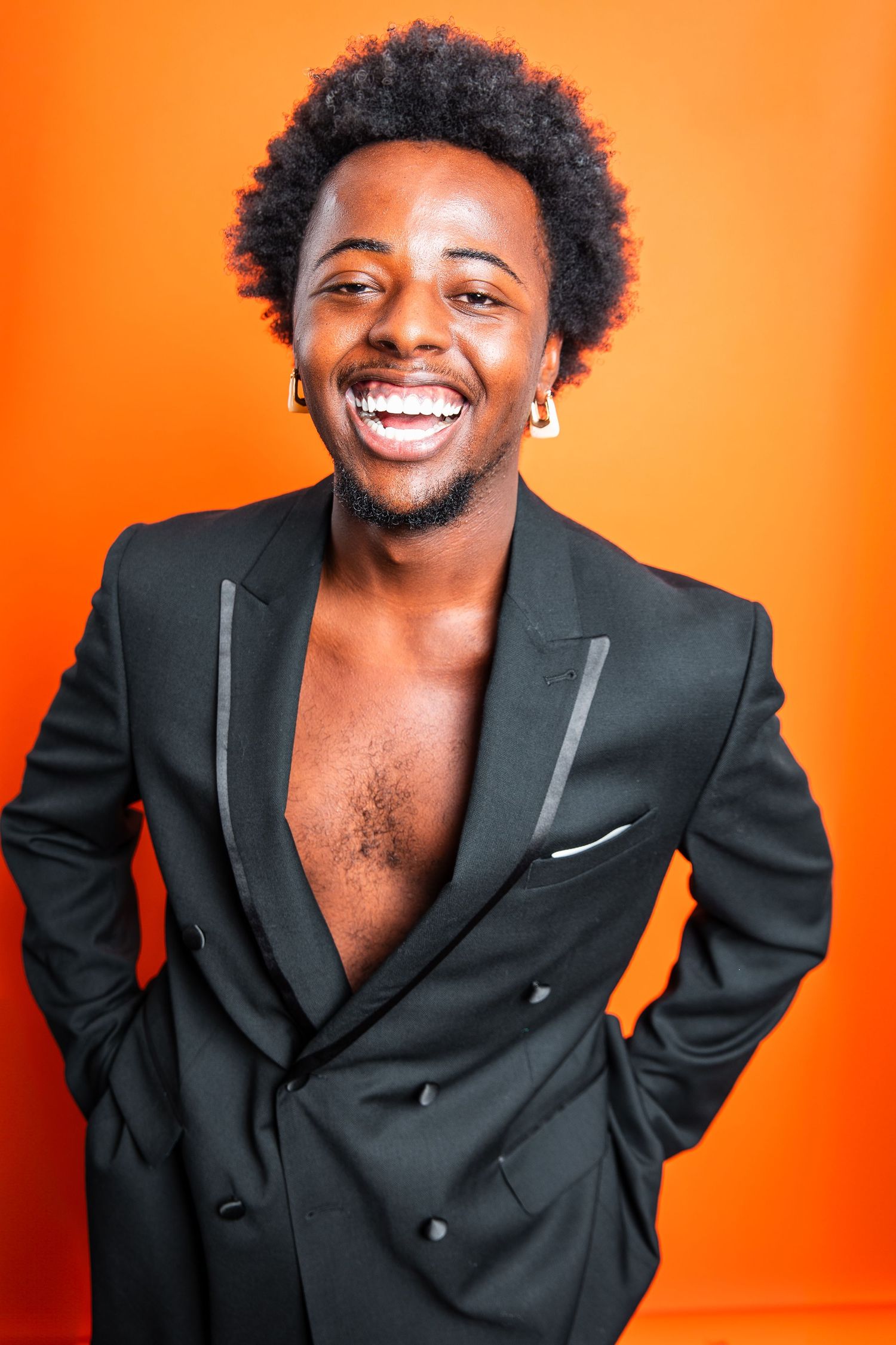 Smiling Black man with afro wearing open black blazer with no shirt against orange background.