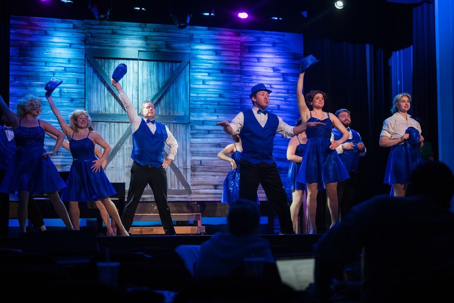 Ensemble cast in blue costumes performs an energetic musical theater number on a rustic stage.