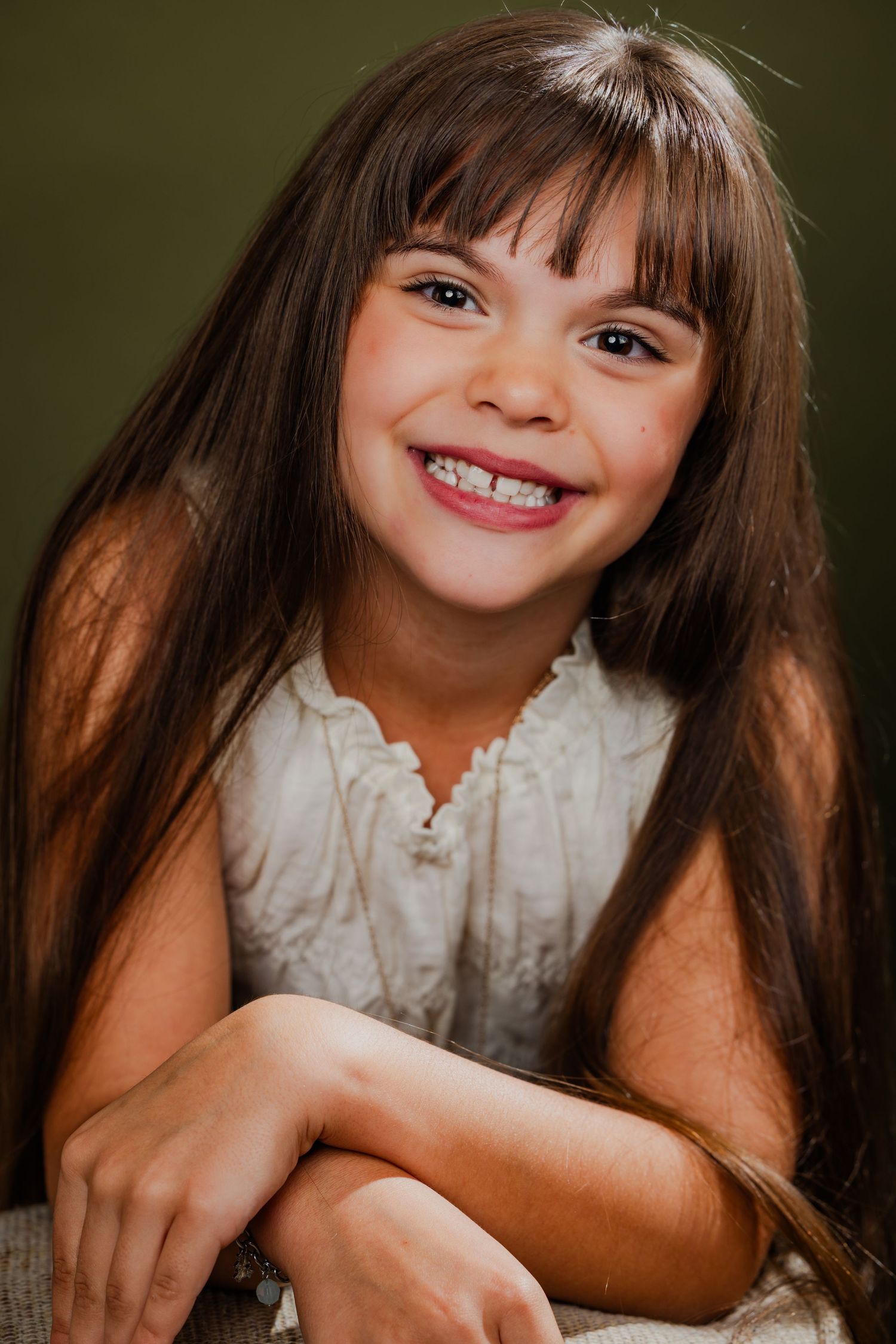 Smiling young girl with long brown hair wearing a white dress seated against a green background.