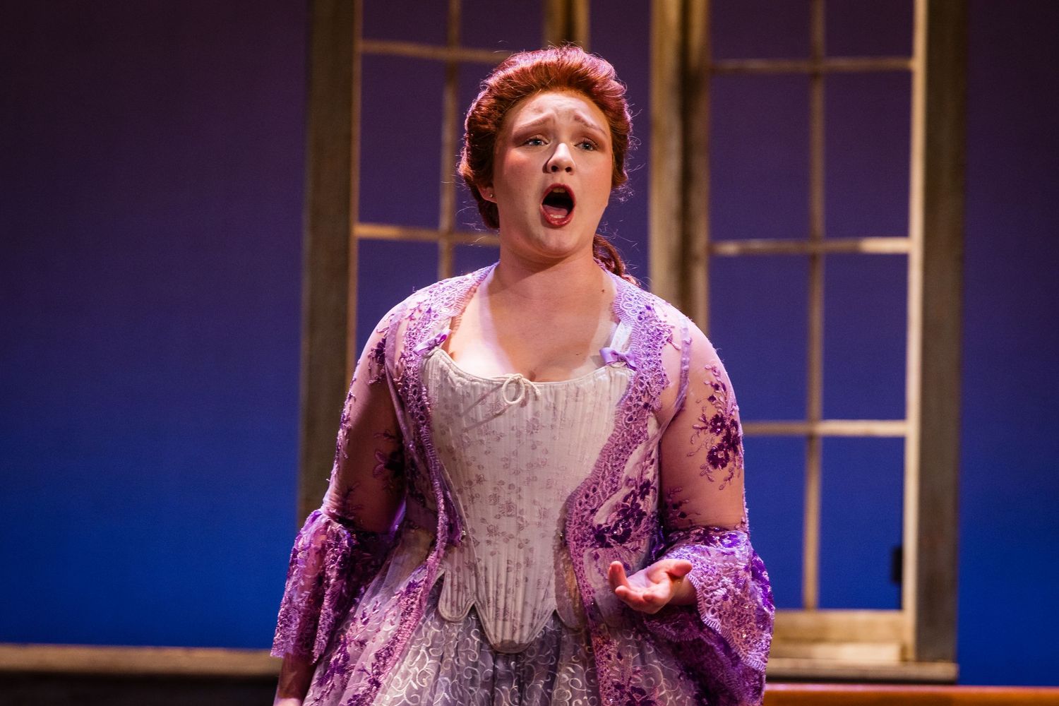 Female opera singer performing on stage in an elegant purple and white period gown.