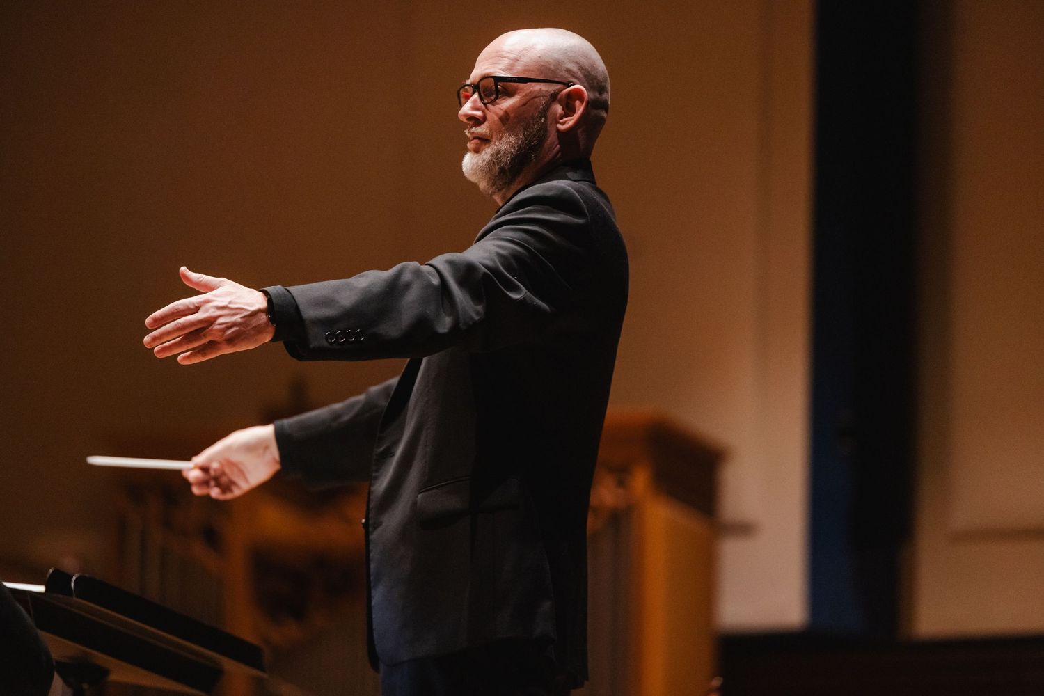 Bearded male conductor in black suit passionately leads orchestra with baton outstretched on stage.
