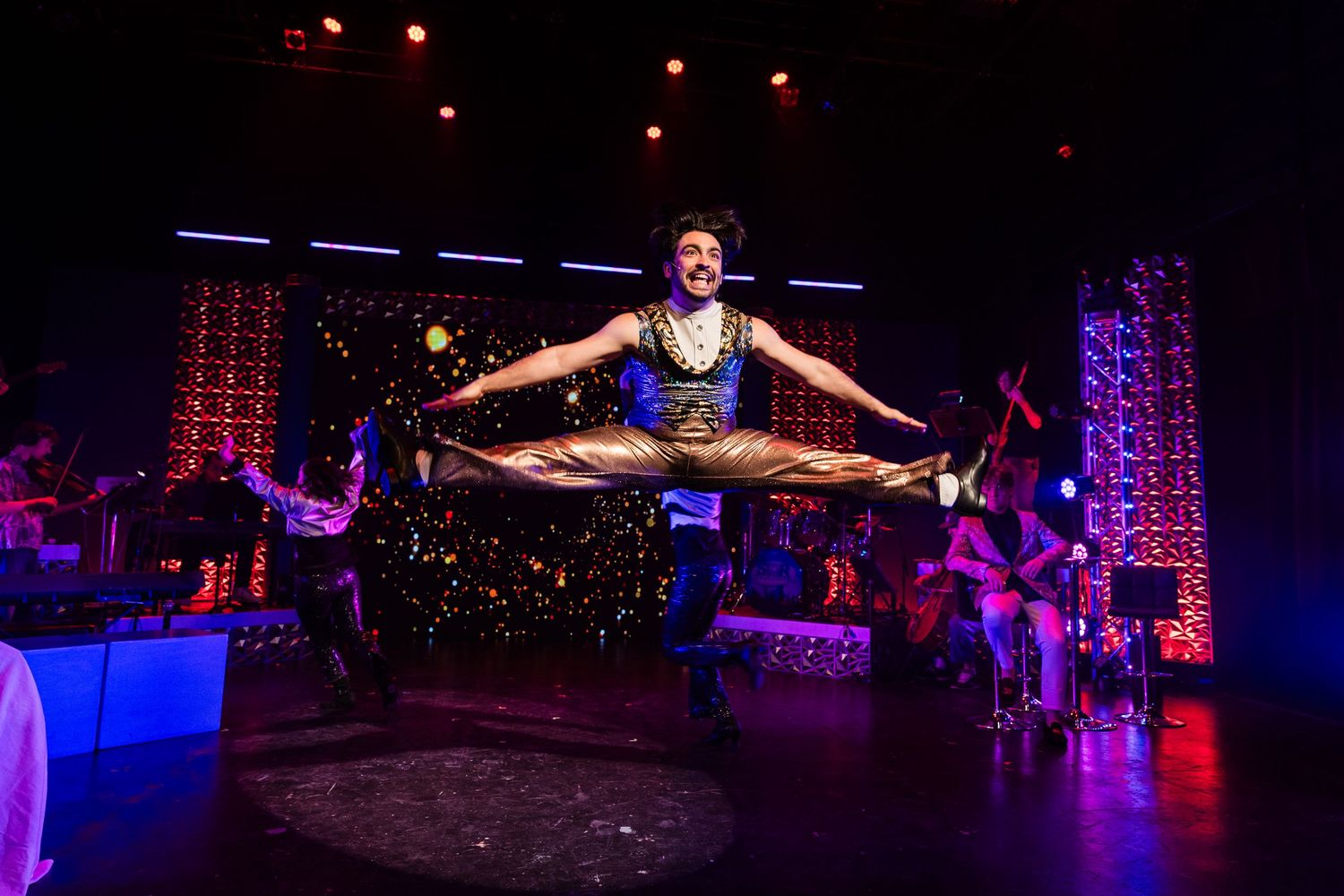 Male performer executes stunning aerial splits jump during vibrant theatrical stage production.