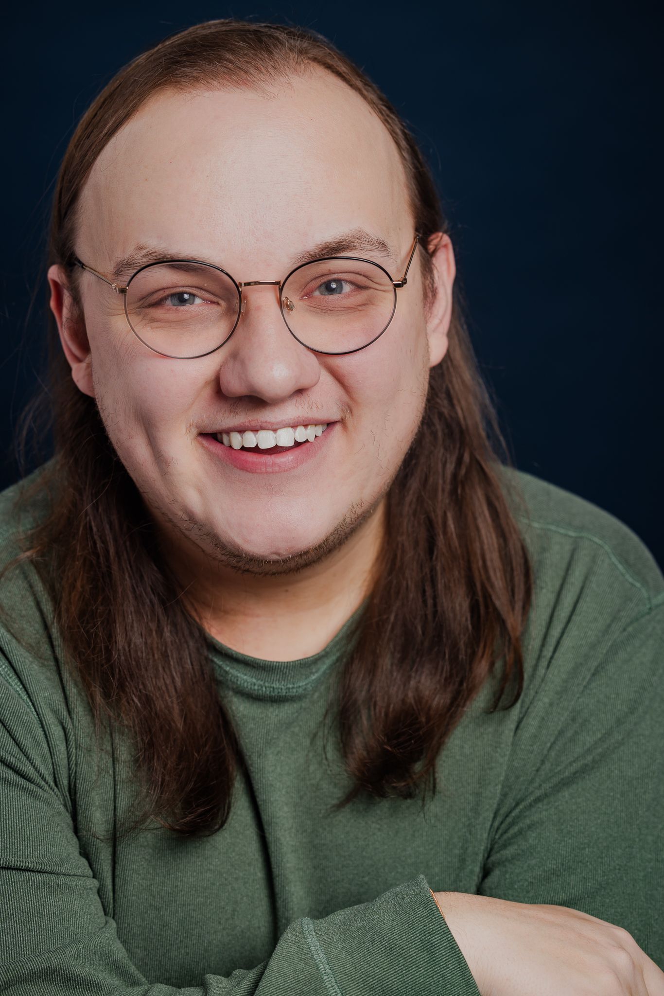 Smiling man with long brown hair and round glasses wearing a green sweater against a dark blue background.