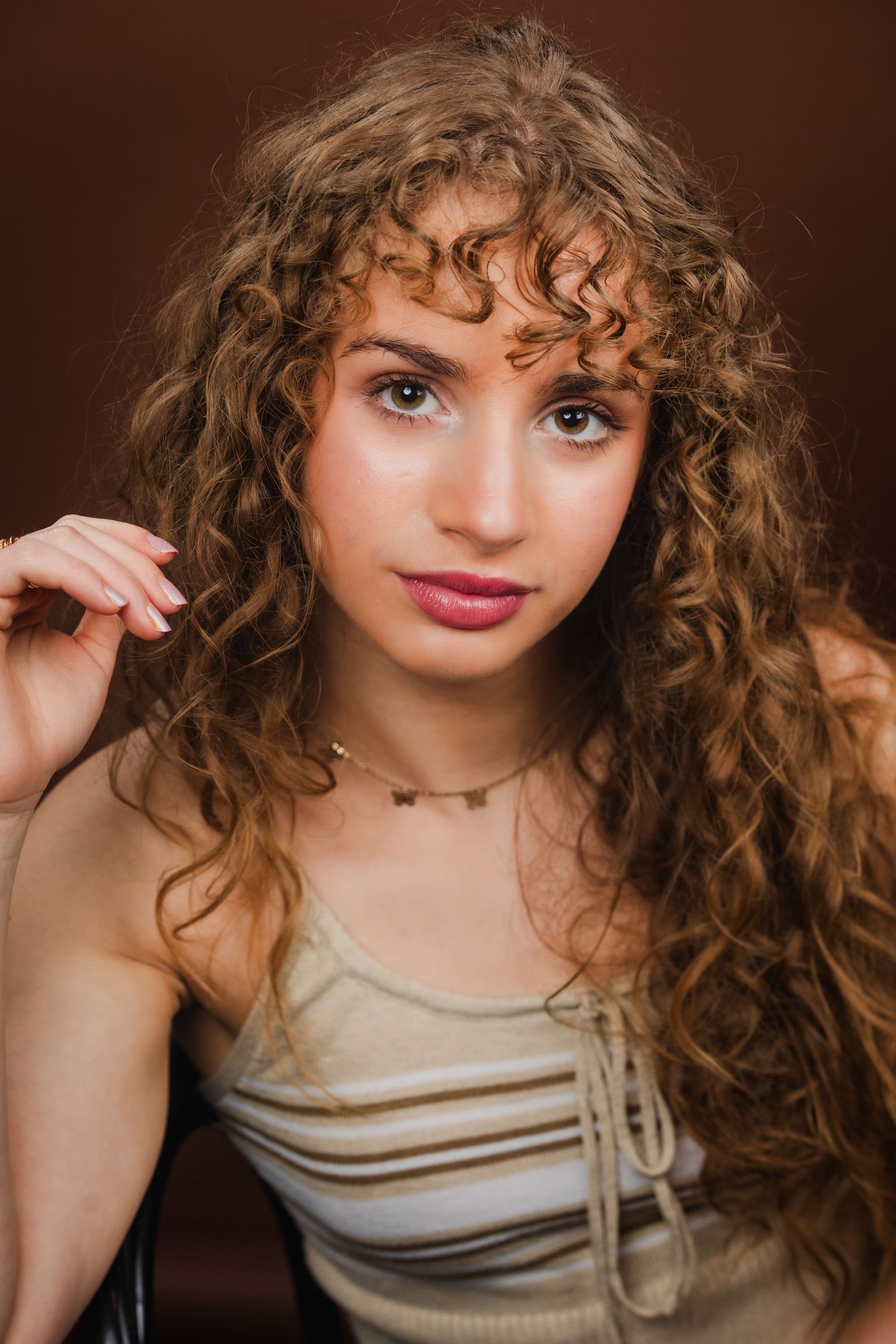 Young woman with long curly auburn hair and hazel eyes wearing a striped top and gold necklace against a brown background.