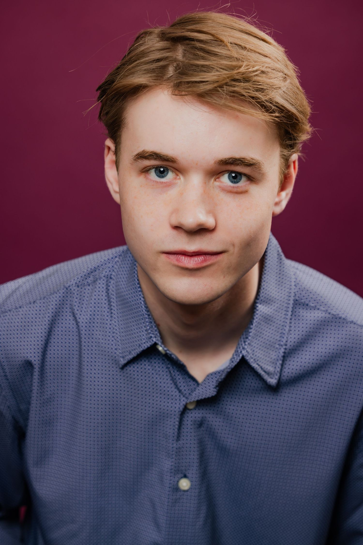 Young man with blonde hair and blue eyes wearing blue button-up shirt, studio portrait.