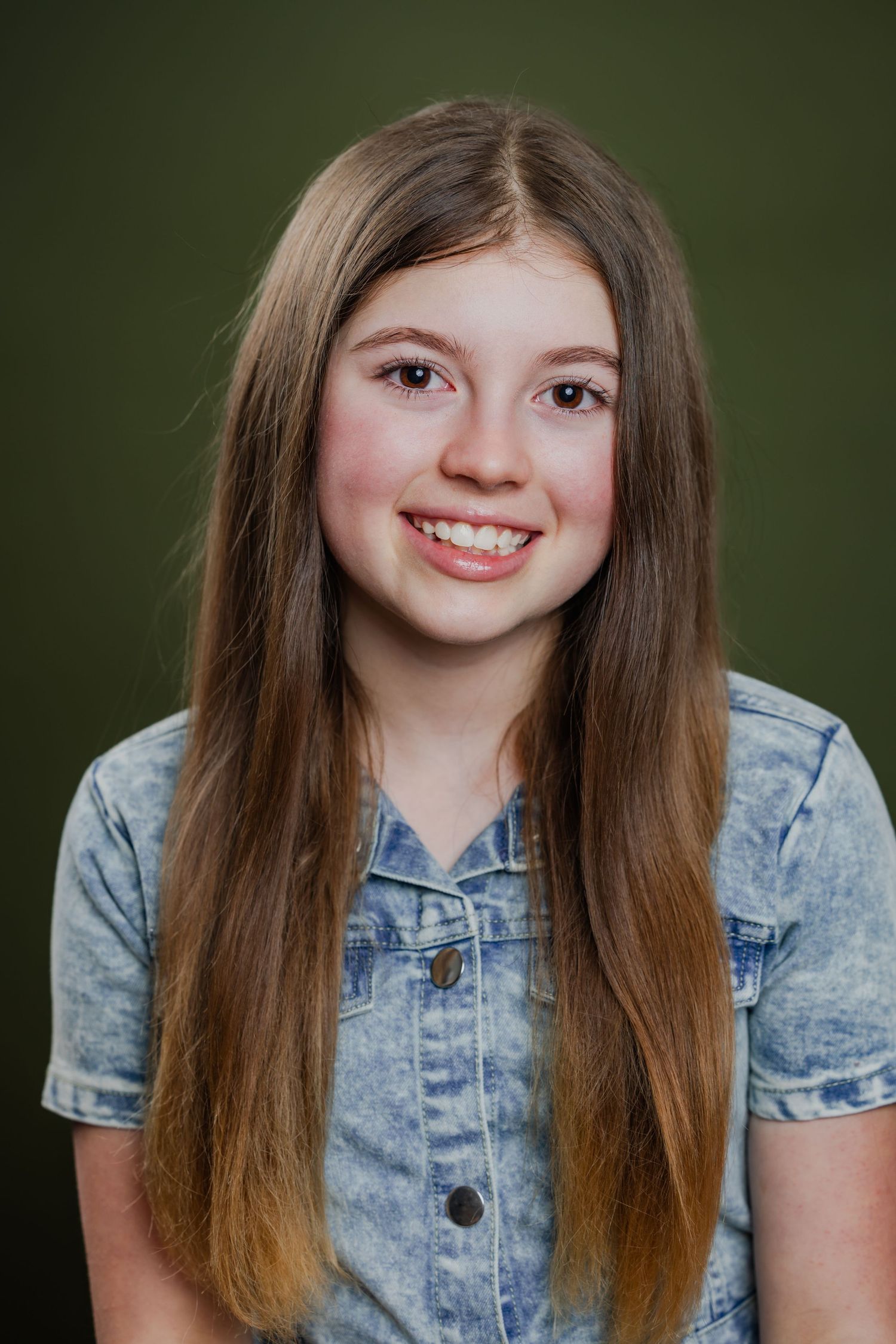 Teenage girl with long brown hair smiling brightly in a denim shirt against a green background.