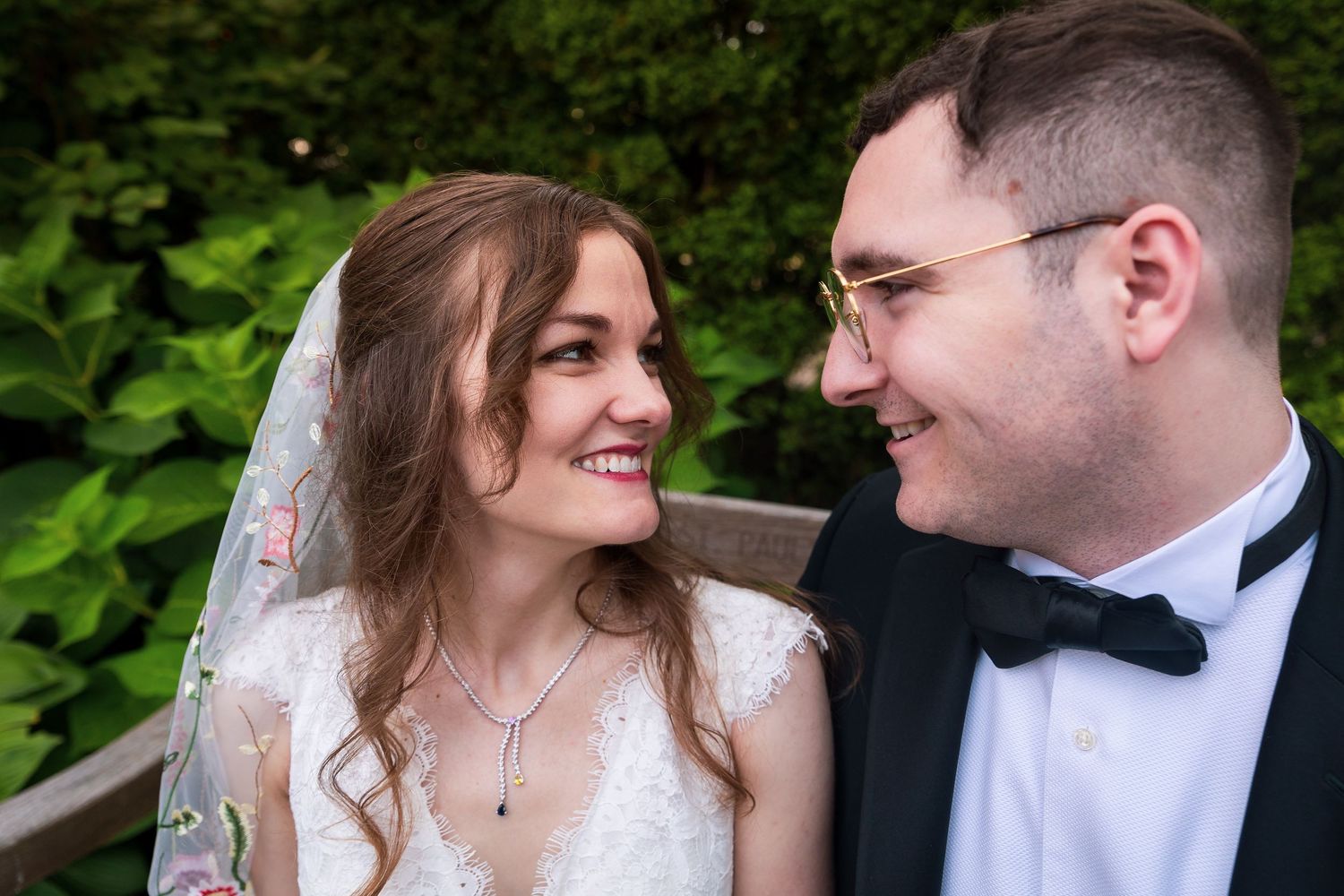Smiling bride and groom in wedding attire gazing at each other with lush green foliage background.