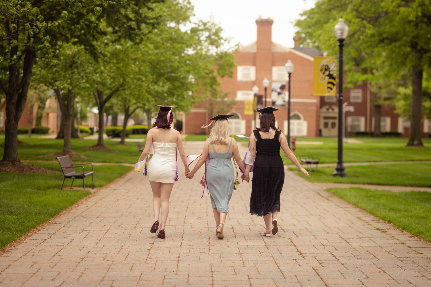 Three female graduates walking hand in hand down a tree-lined campus brick pathway.