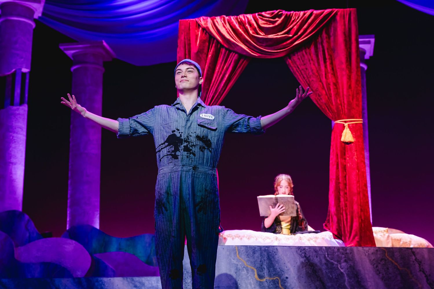 A performer in blue overalls stands with arms outstretched on a theatrical stage with red velvet curtains.
