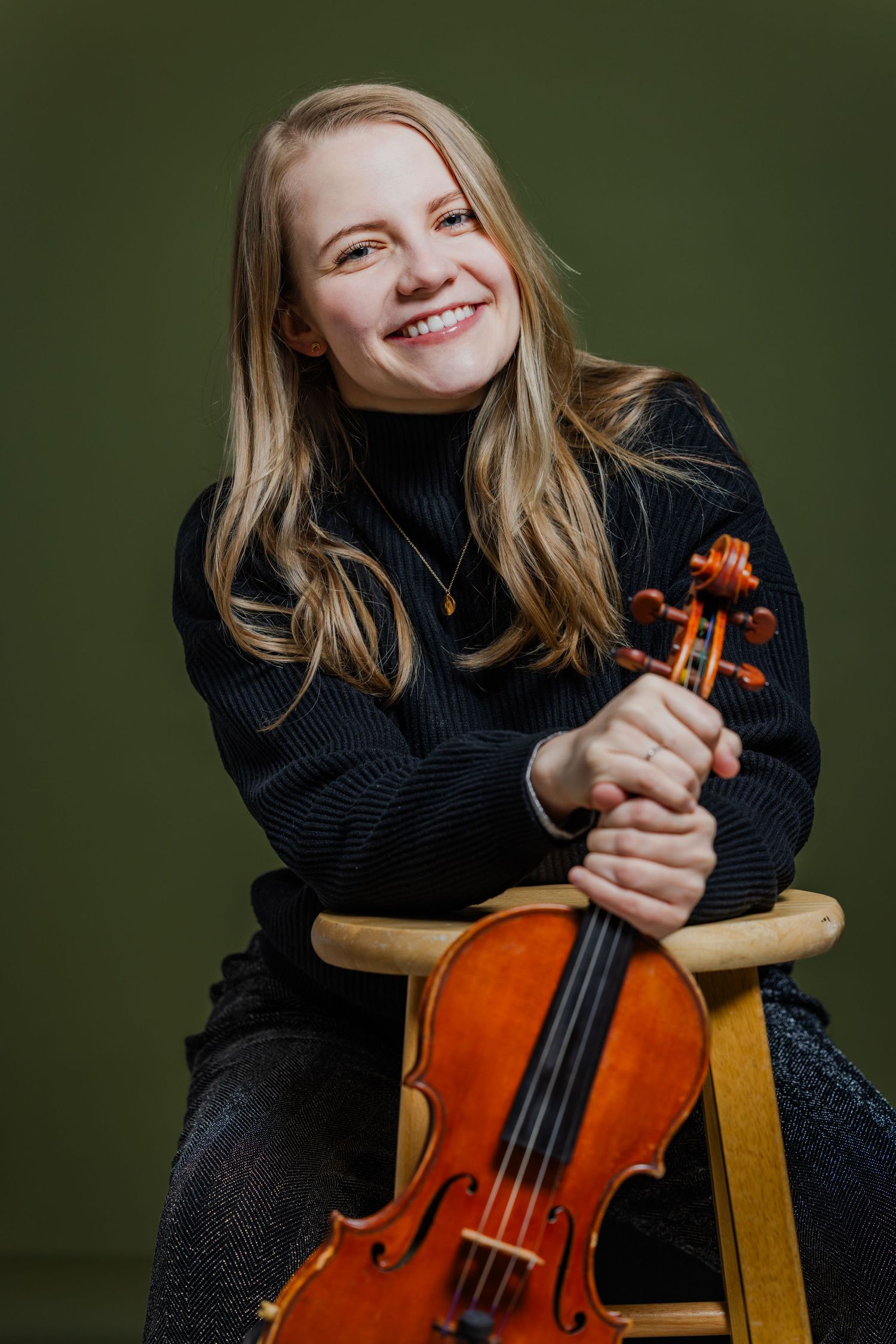 Smiling blonde violinist sitting with her violin, wearing a black turtleneck sweater on a green background.