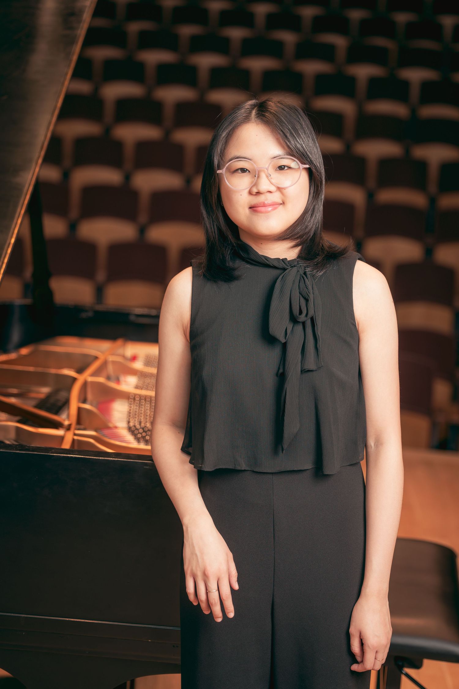 Young woman with glasses standing beside a grand piano in a concert hall.