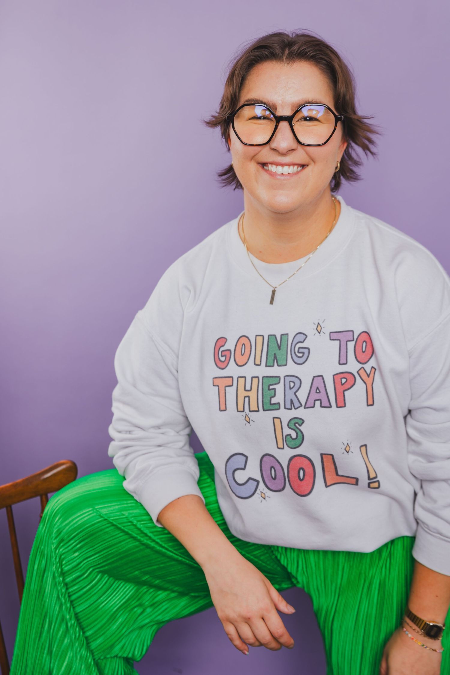 Smiling woman in glasses wearing therapy sweatshirt and green pants seated against purple background.