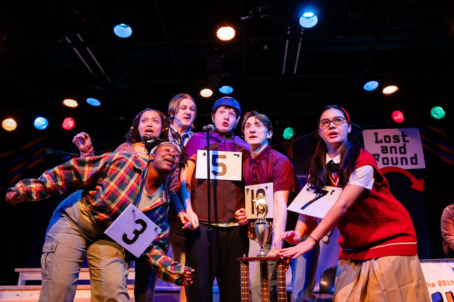 Five young performers on stage in a colorful spelling bee musical theater production wearing numbered contestant badges.