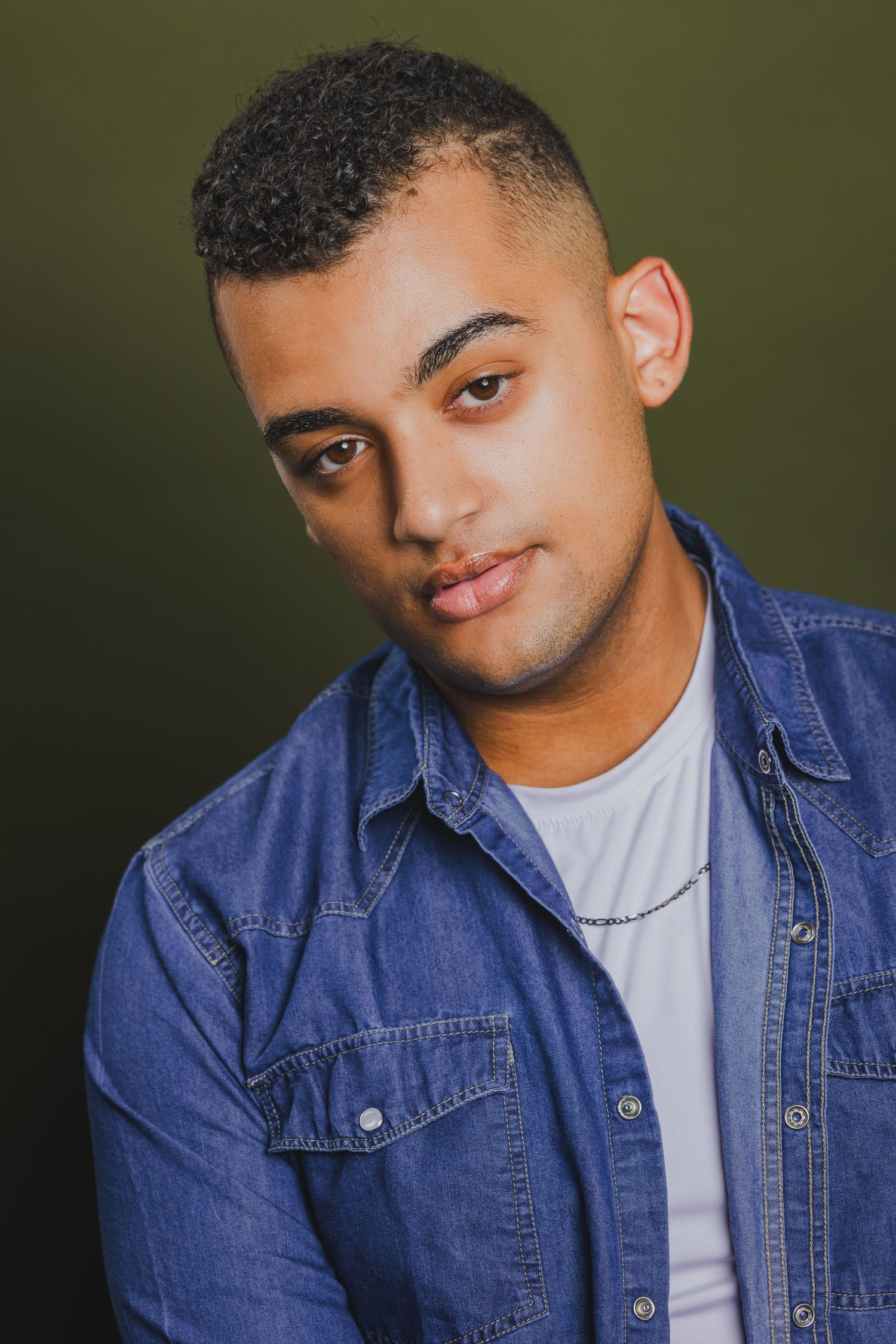 Young man with short curly hair wearing denim shirt and silver chain necklace against dark green background.