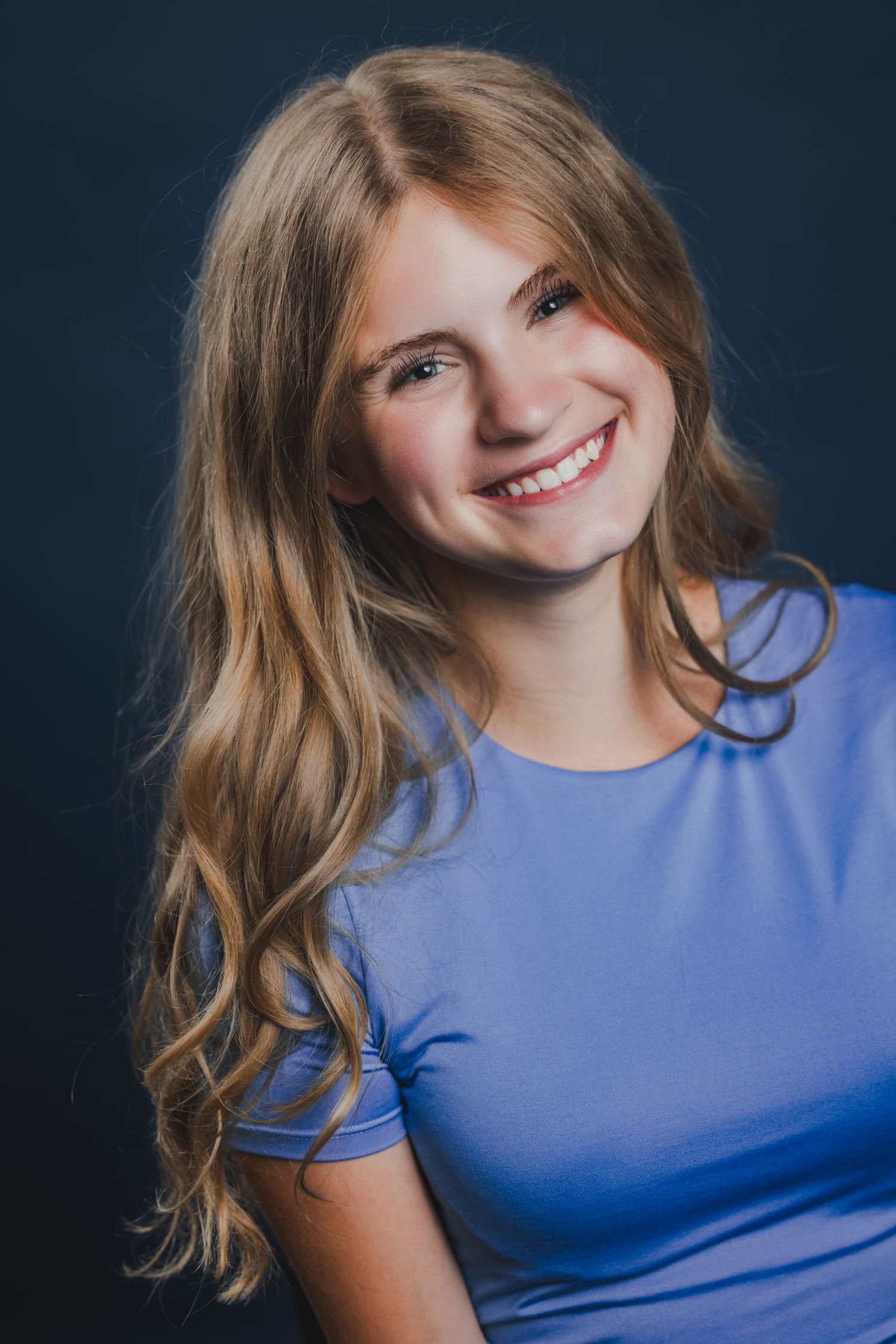 Cheerful teenage girl with long blonde wavy hair wearing a lavender t-shirt, smiling against a dark studio background.