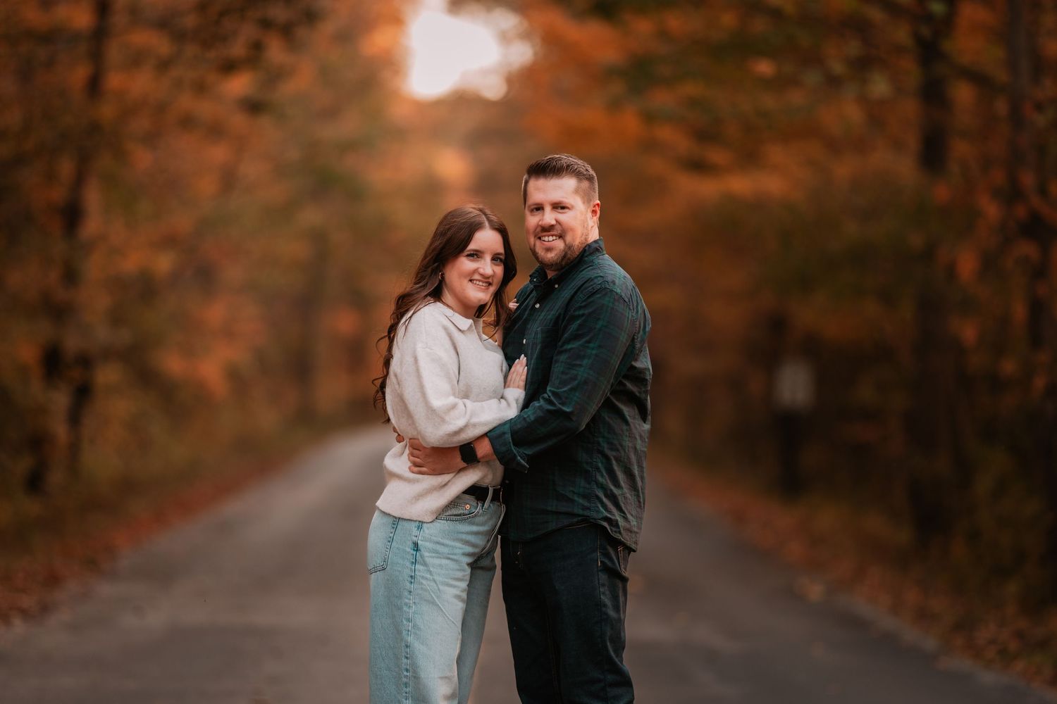 A happy couple embraces on a tree-lined road surrounded by vibrant orange fall foliage.