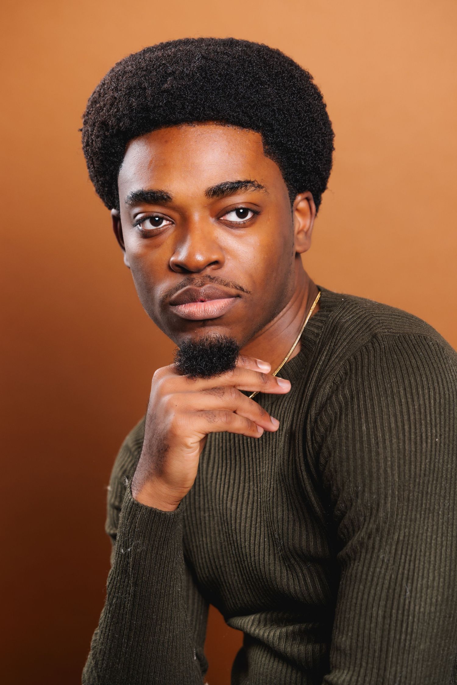 Young Black man with afro resting chin on hand wearing olive sweater against brown background.