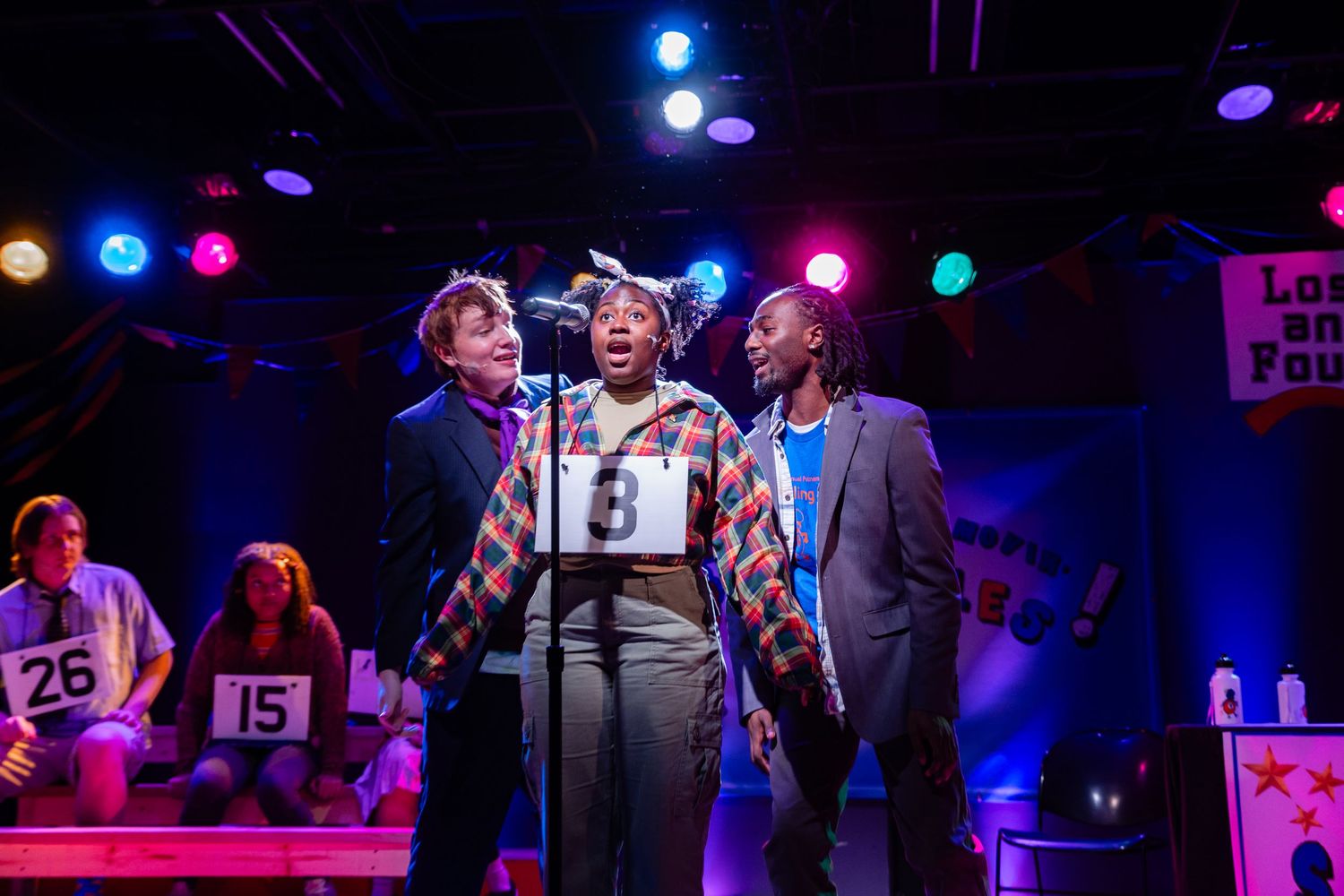 Three theatrical performers singing together on a colorful stage during a spelling bee musical production.