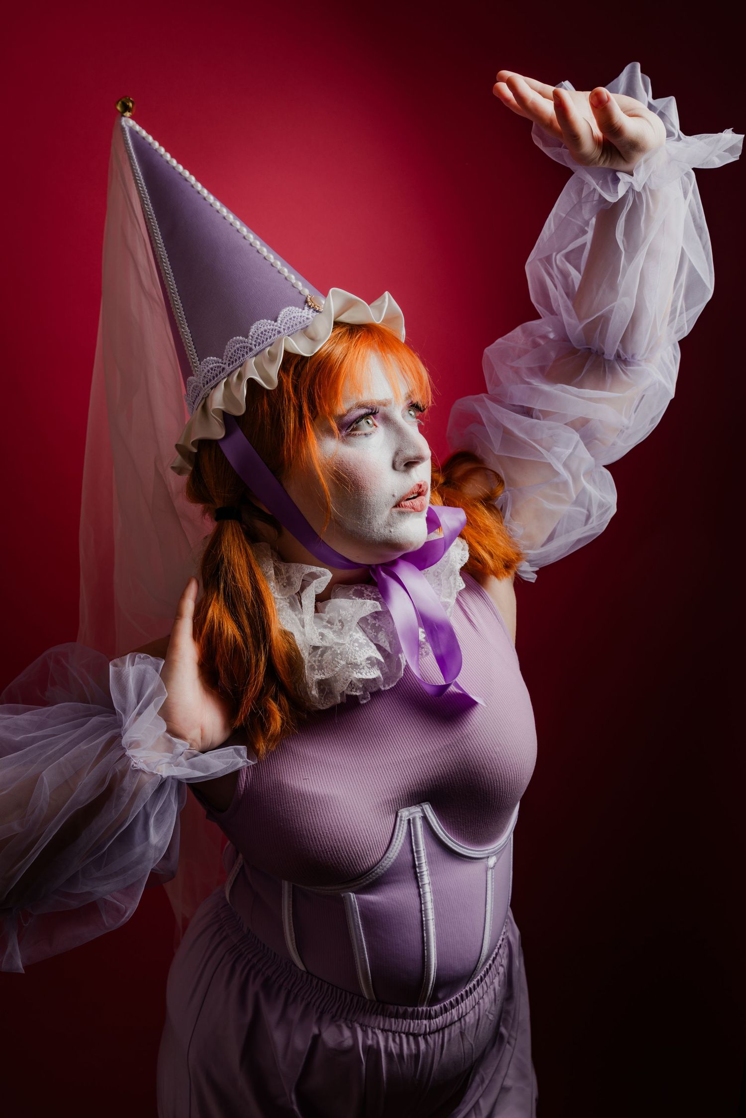 Woman in purple fantasy costume with cone hat, white face paint, and orange hair against a red background.