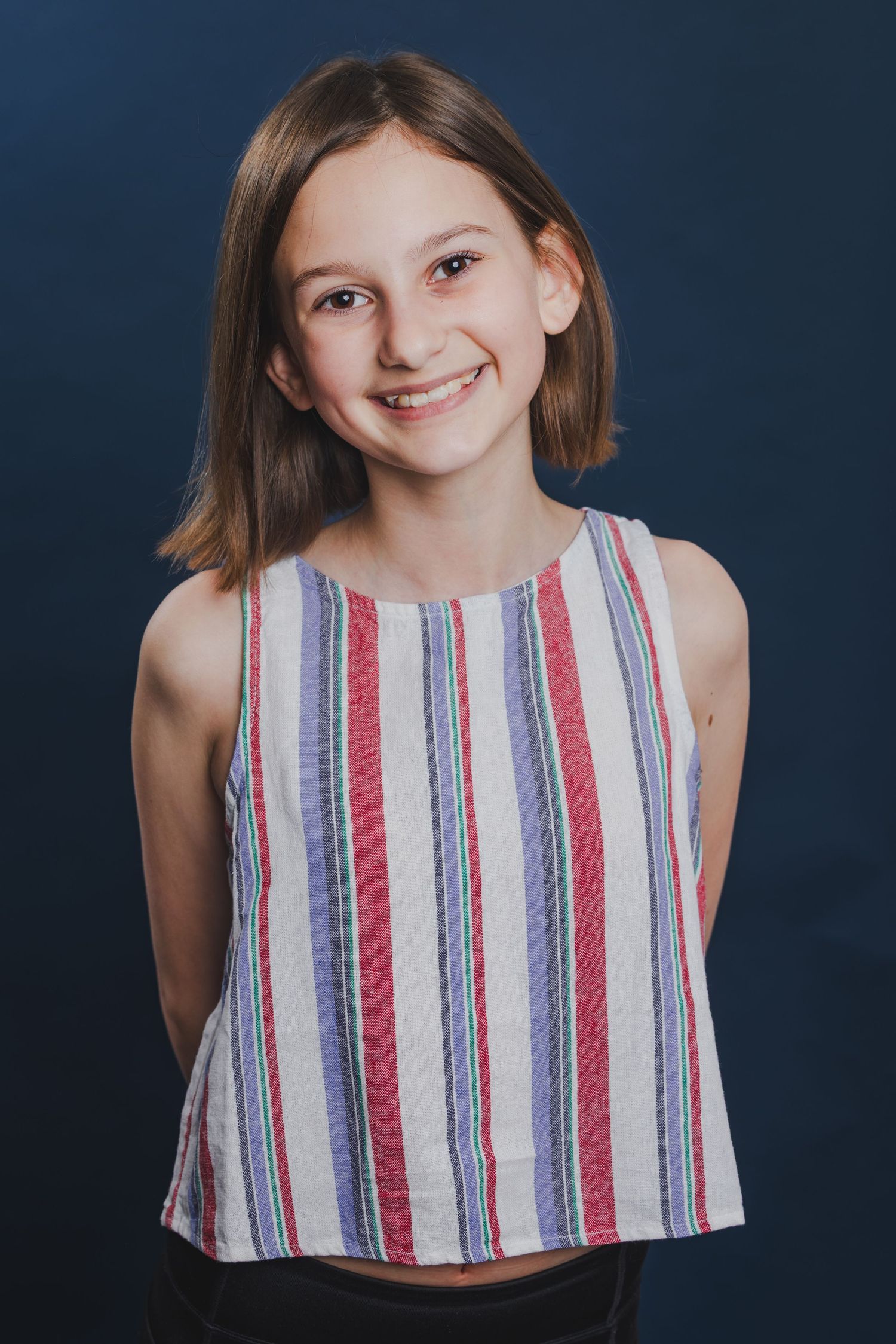 Young girl with brown hair smiling in a striped sleeveless top against a dark blue background.