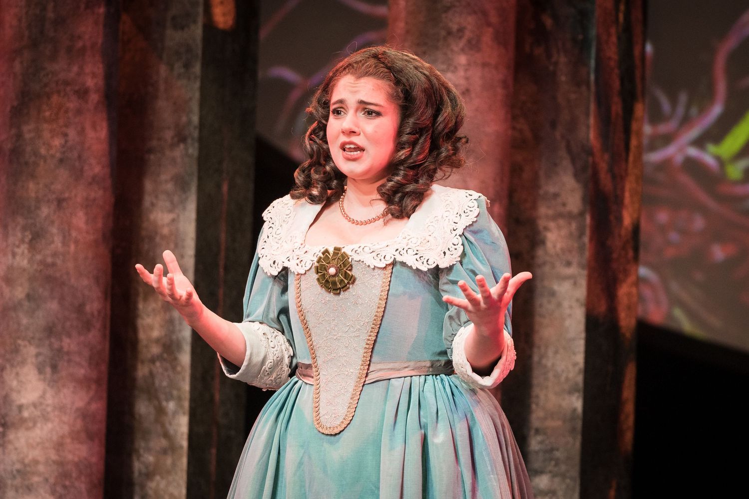 Young woman performer in a teal period costume with white lace collar singing on a theatrical stage.