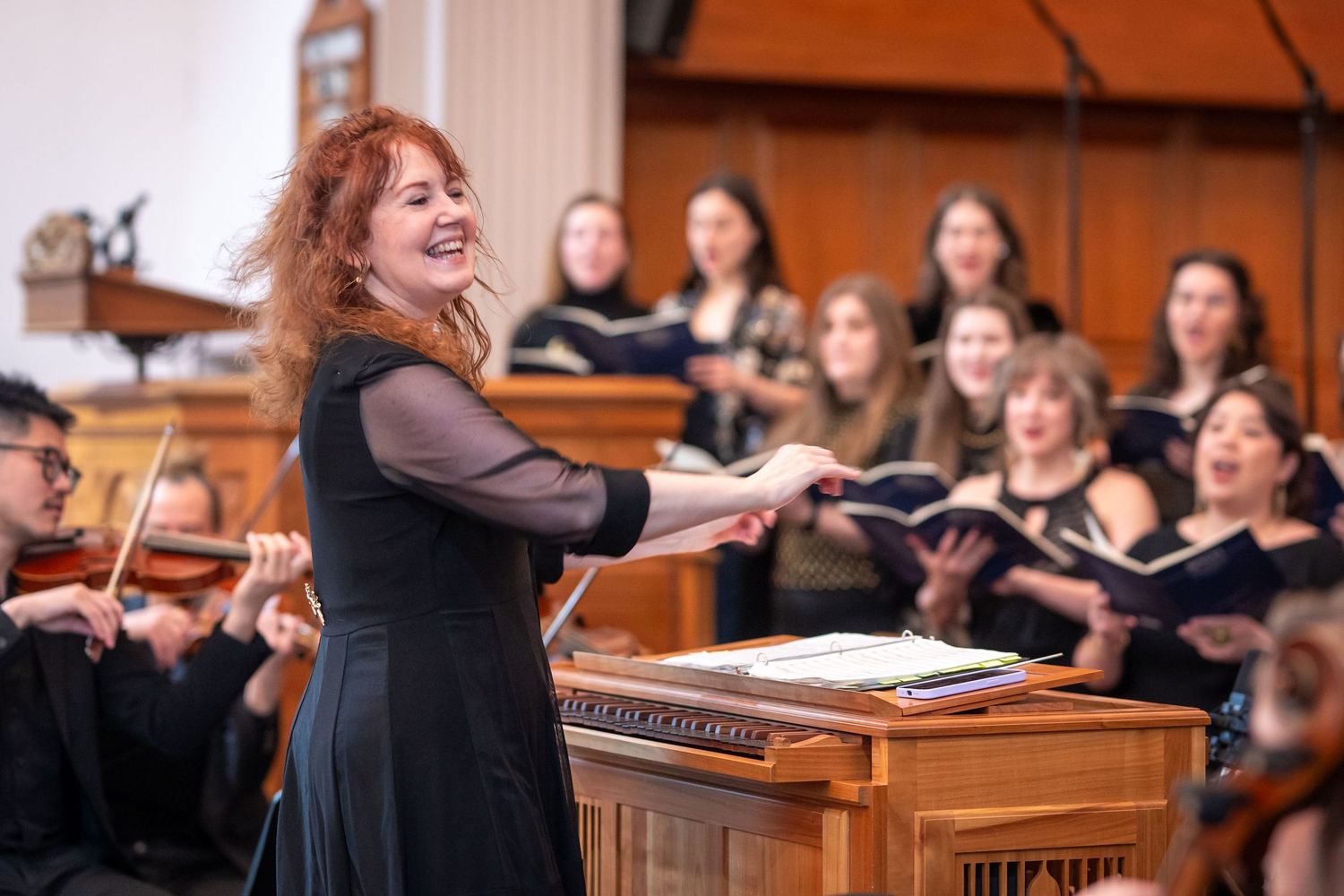 A smiling red-haired conductor gestures warmly before a choir of young women in a church.