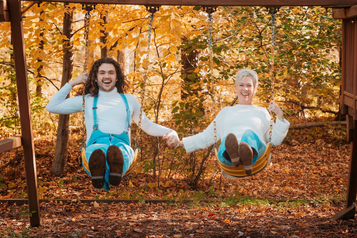 Couple holding hands on swings in autumn forest with golden fall foliage background.