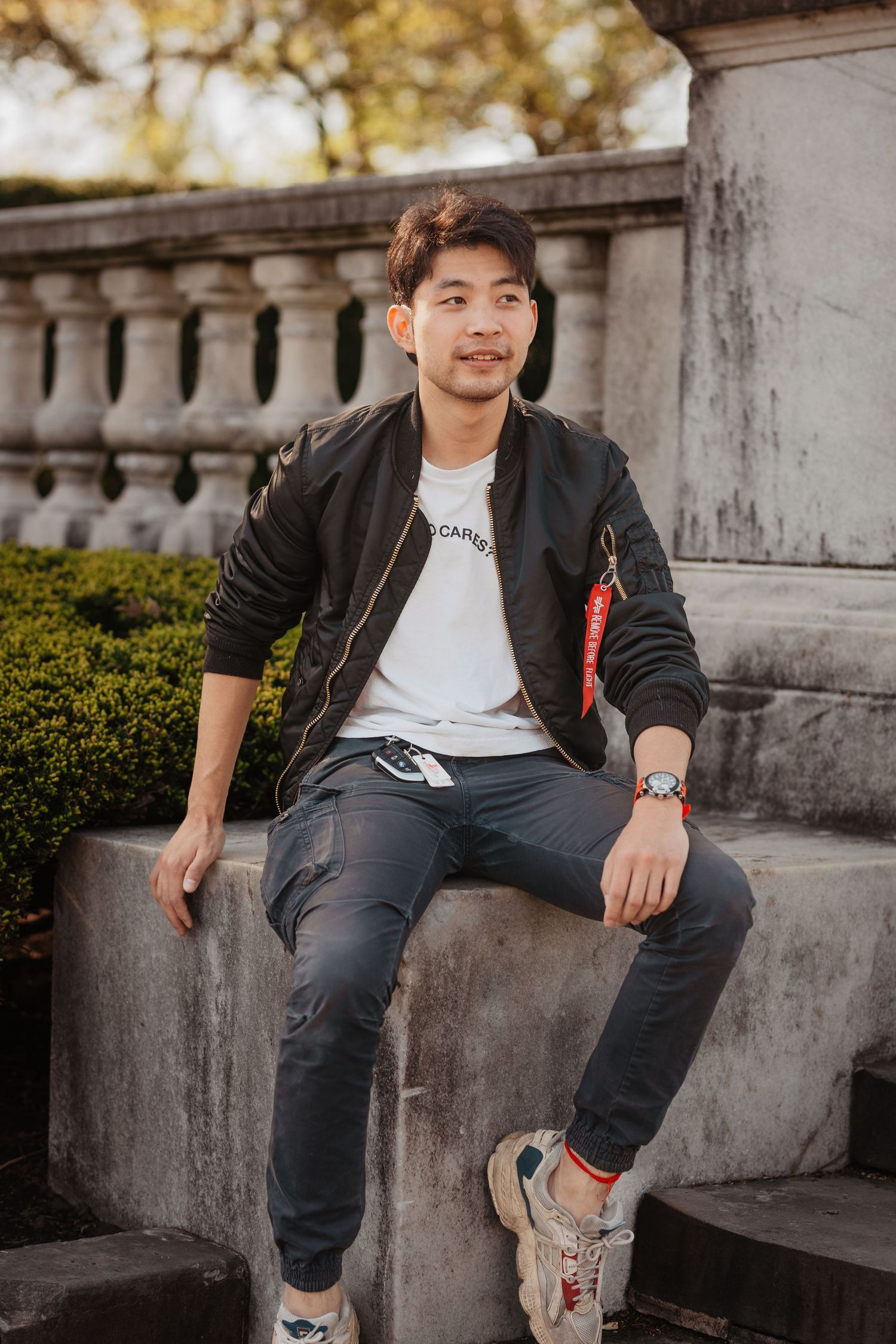 Young Asian man in black bomber jacket sitting on stone steps in an outdoor urban setting.
