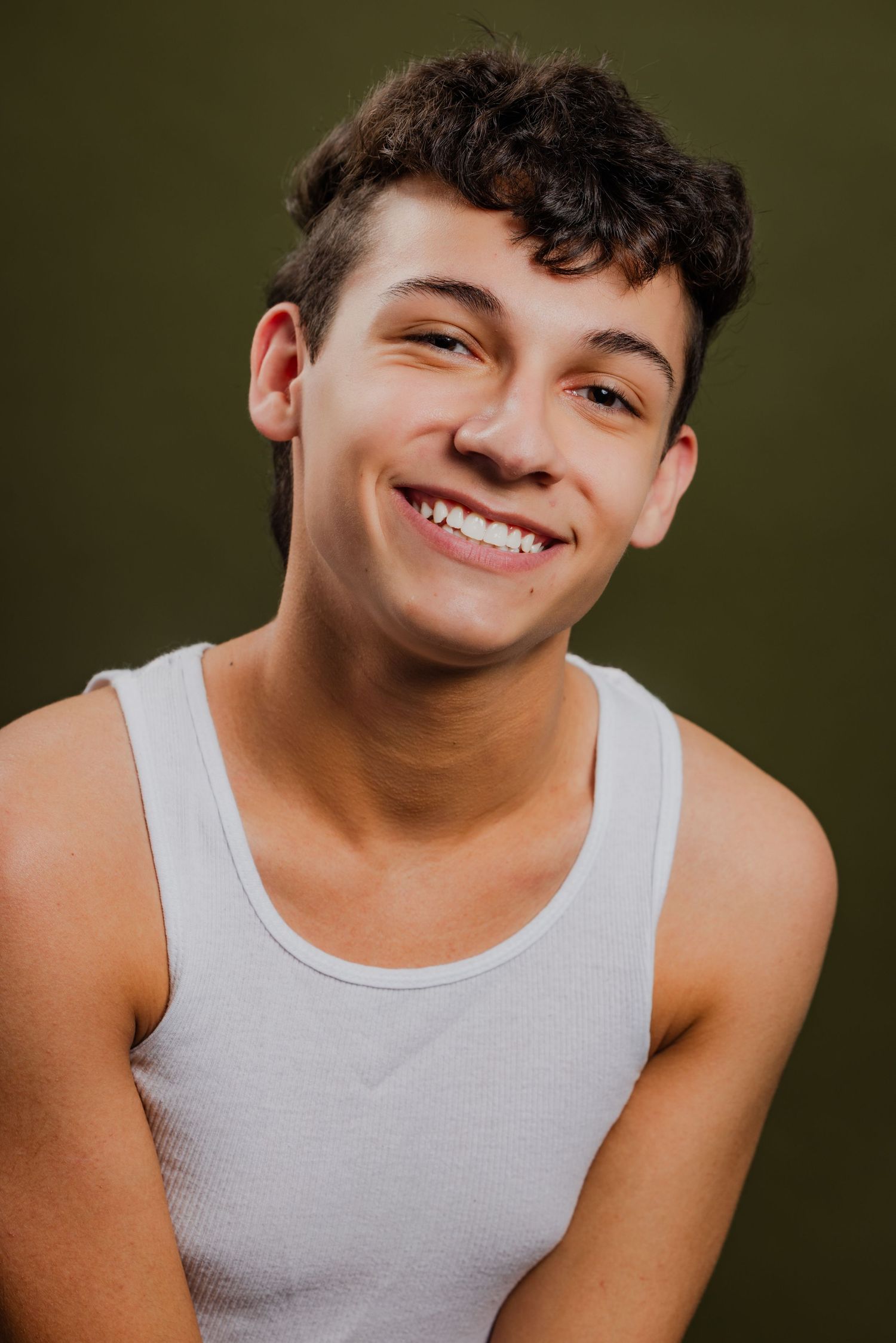 Happy teenage boy with curly brown hair wearing a white tank top smiling against an olive green background.
