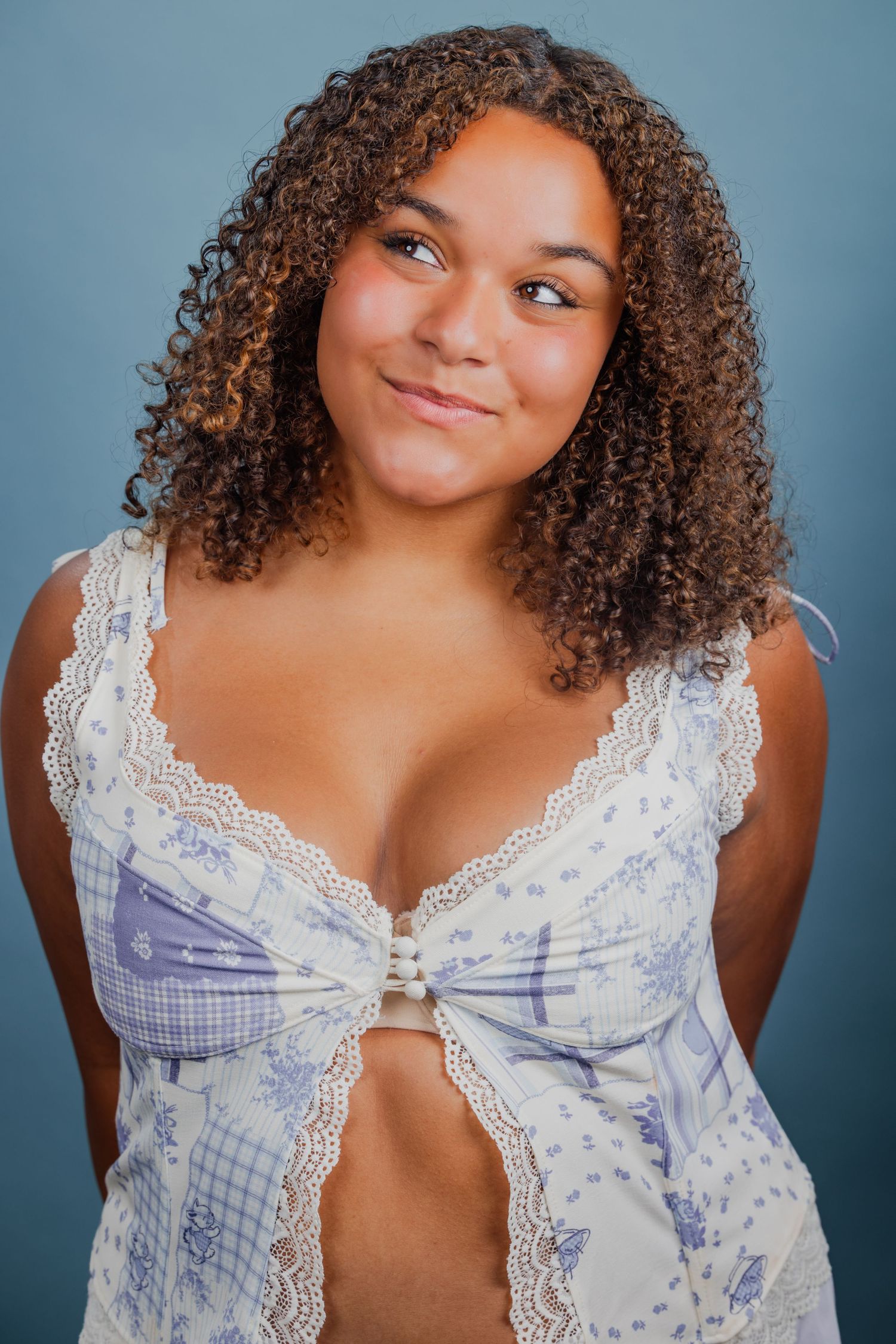 Young woman with curly hair wearing a floral blue and white top against a gray background.