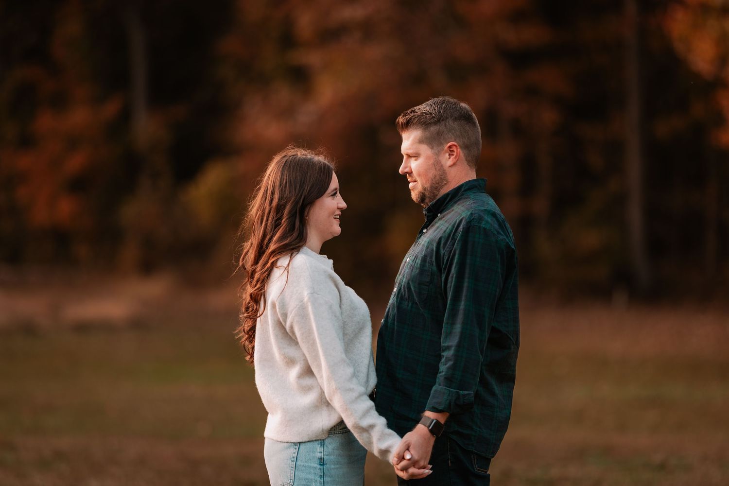 An engaged couple holds hands and gazes at each other in a golden autumn park setting.