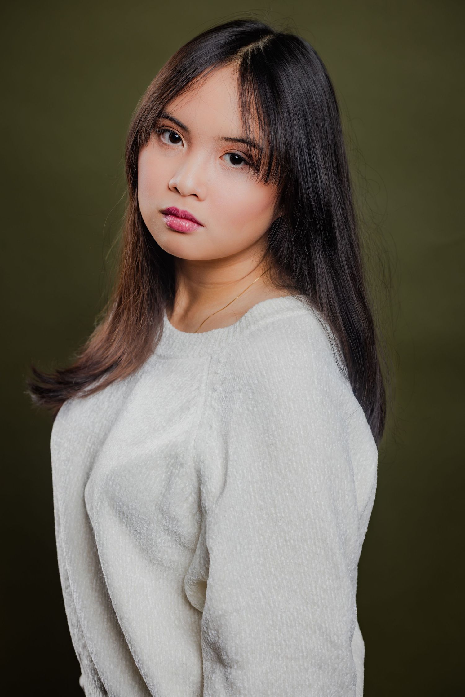 Young Asian woman with long dark hair in white sweater posing against an olive green background.