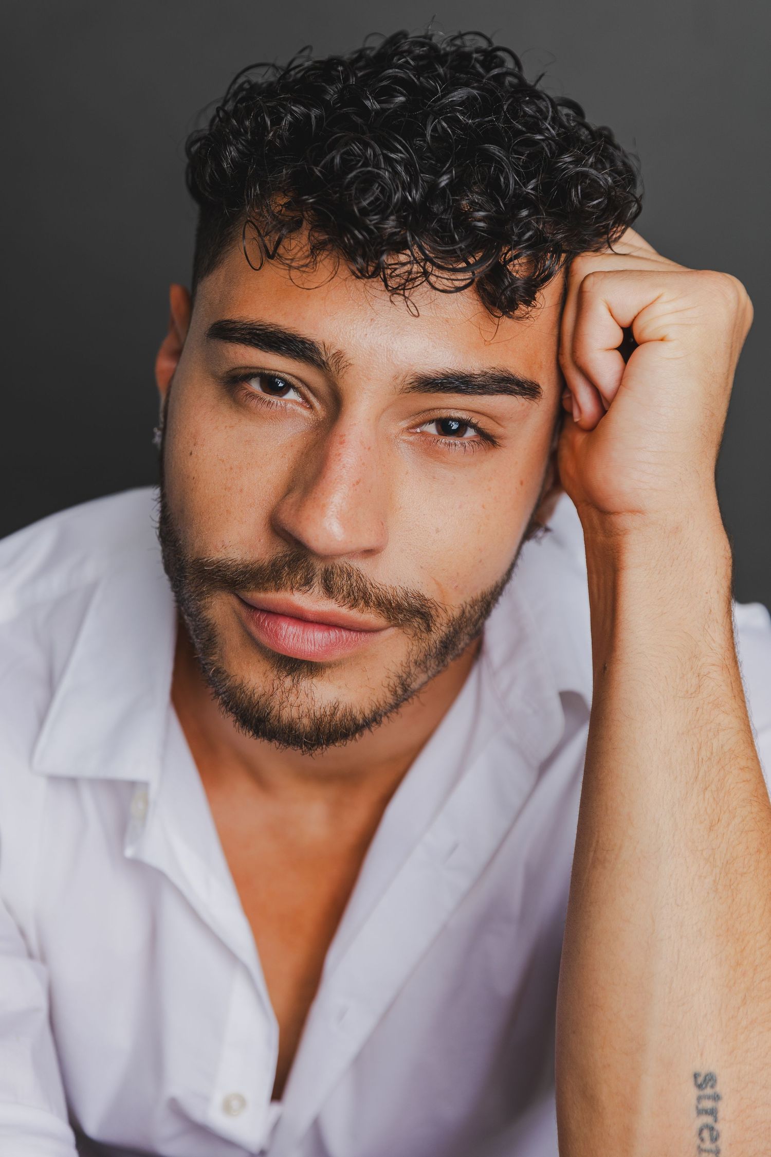 Young man with curly dark hair resting head on hand in white shirt studio portrait.