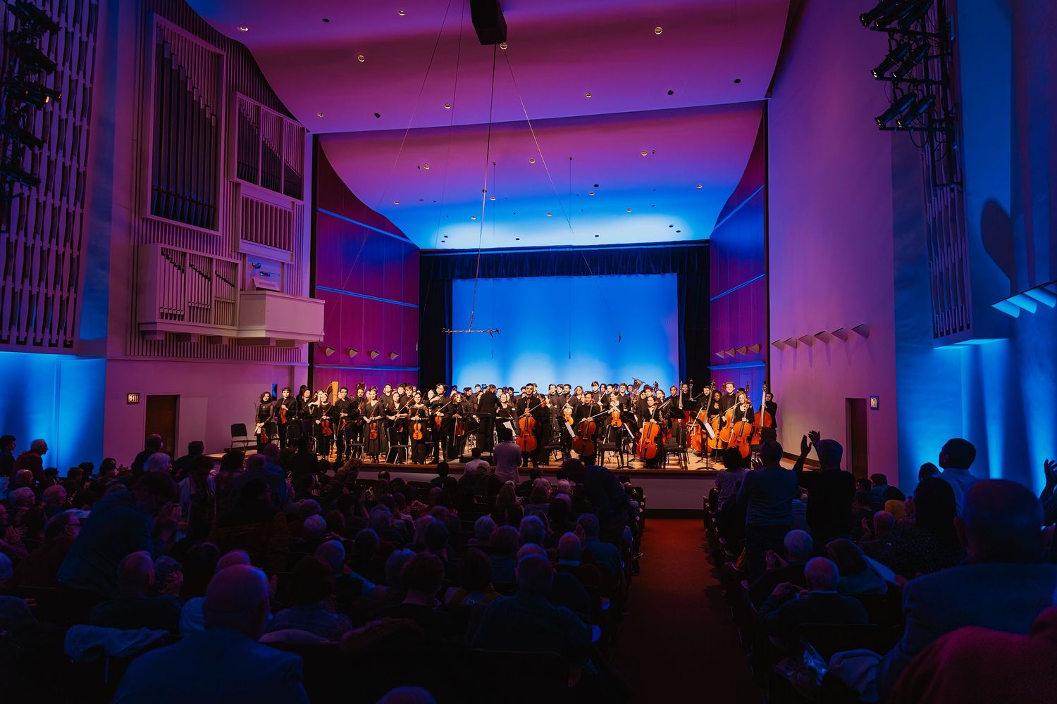 A full orchestra and choir perform on a dramatically lit blue and purple stage in a concert hall.