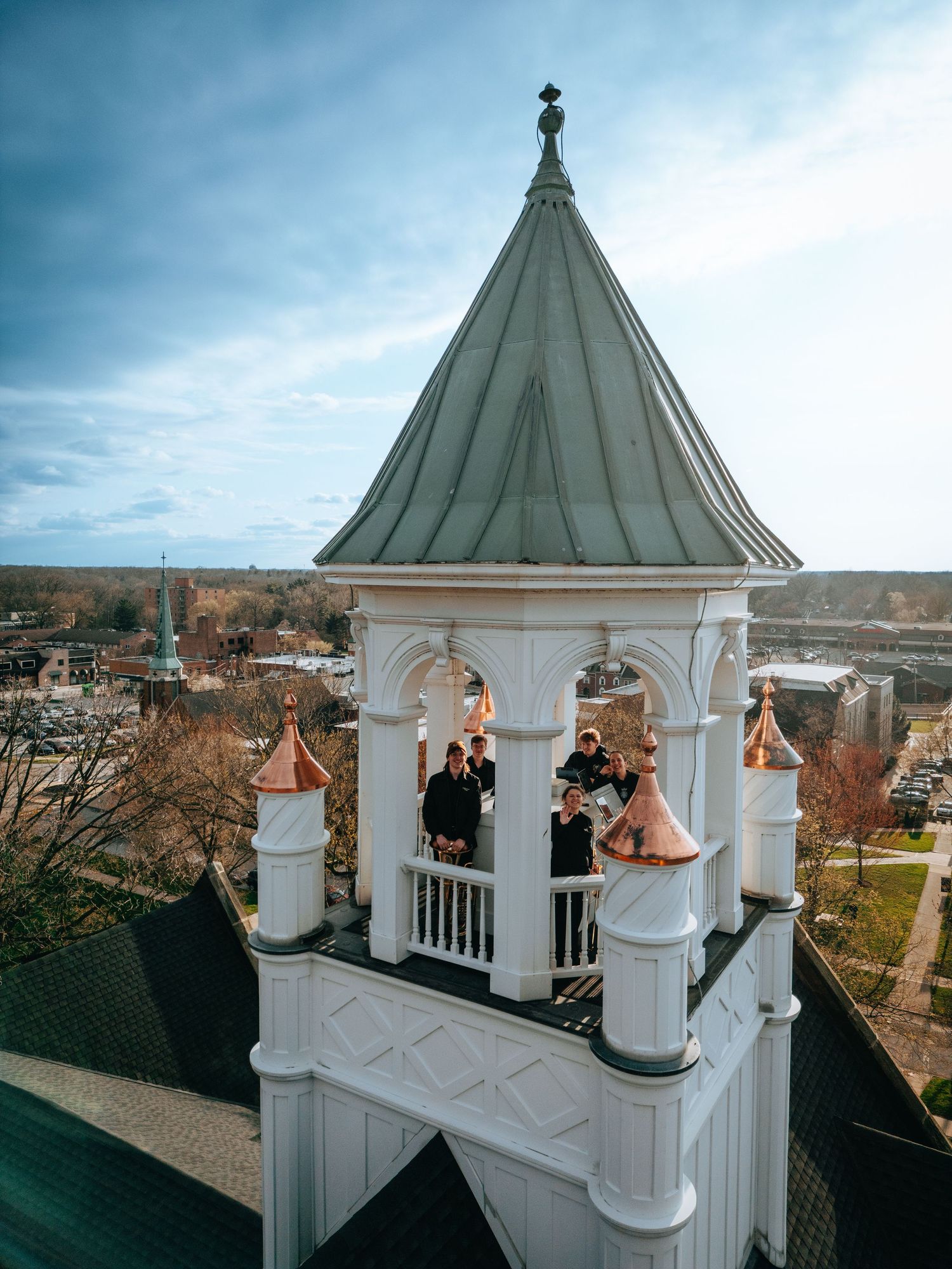 Aerial drone view of people standing on a white church bell tower with copper roof against a cloudy sky.