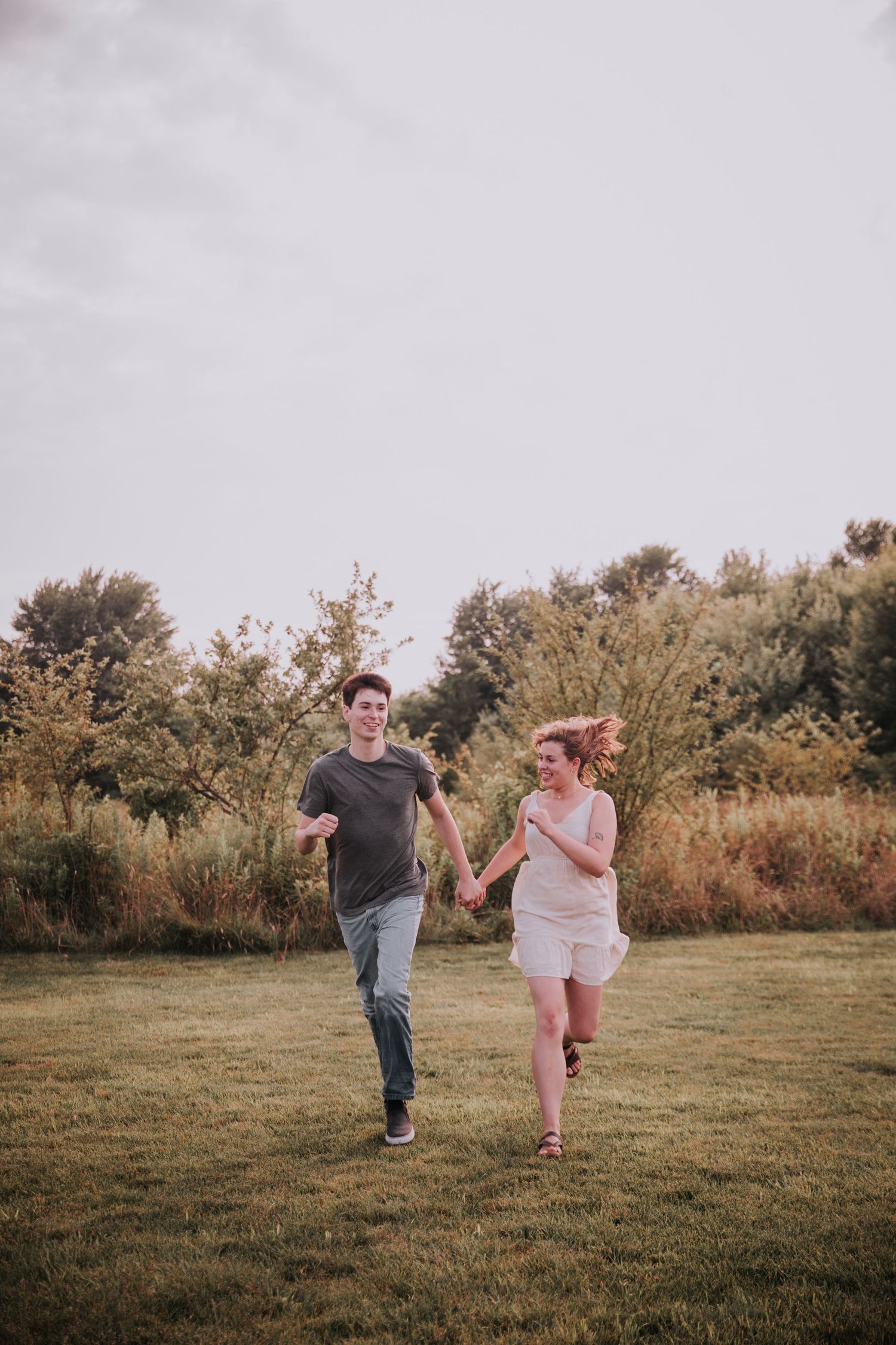 A joyful couple runs hand-in-hand through a lush green field with trees in the background.