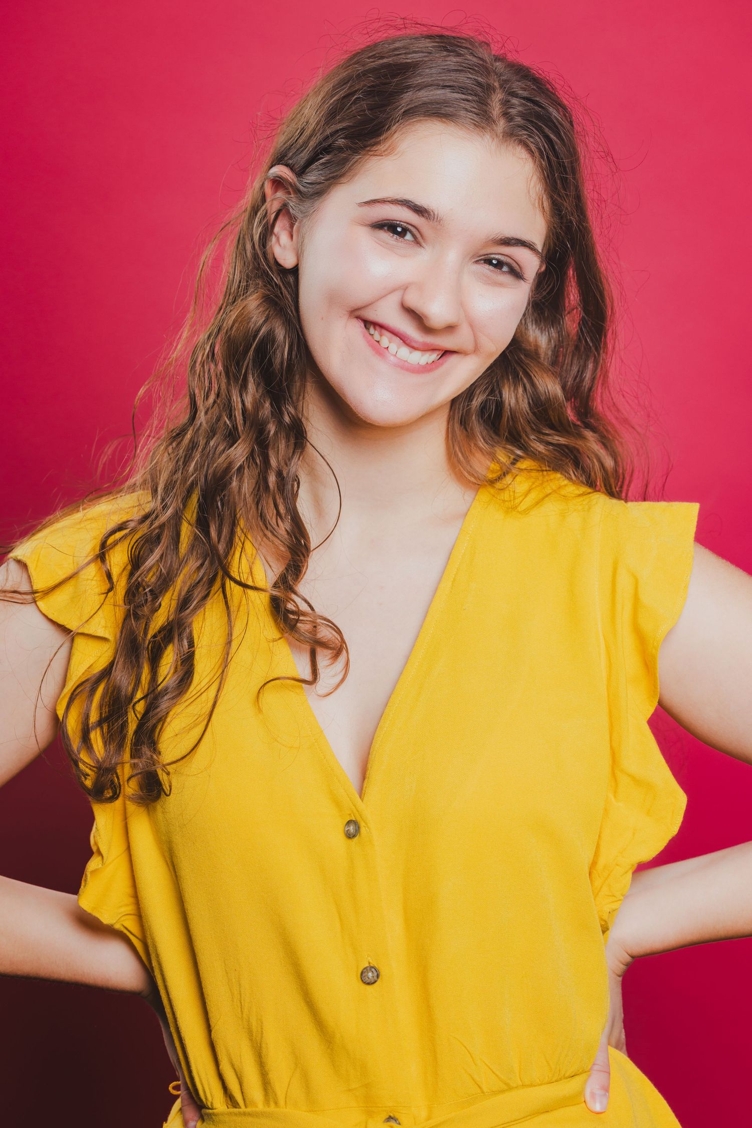 Happy young woman with curly brown hair wearing a yellow button-front dress, posing against a vibrant red background.