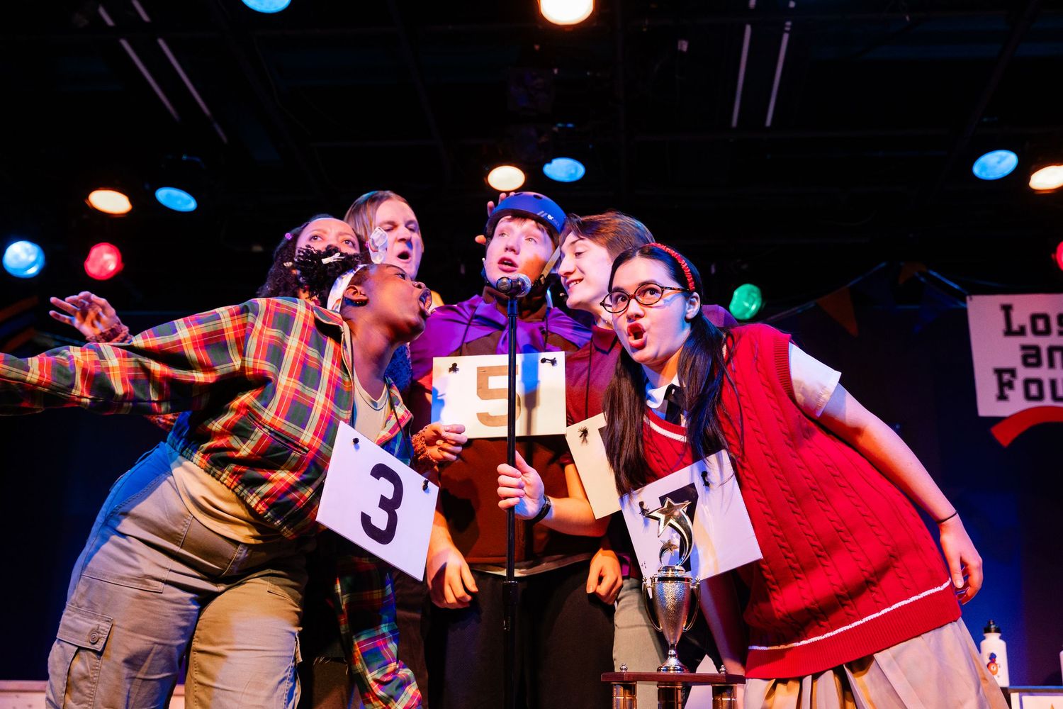 Four energetic performers crowd around a score card in a colorful spelling bee musical production.