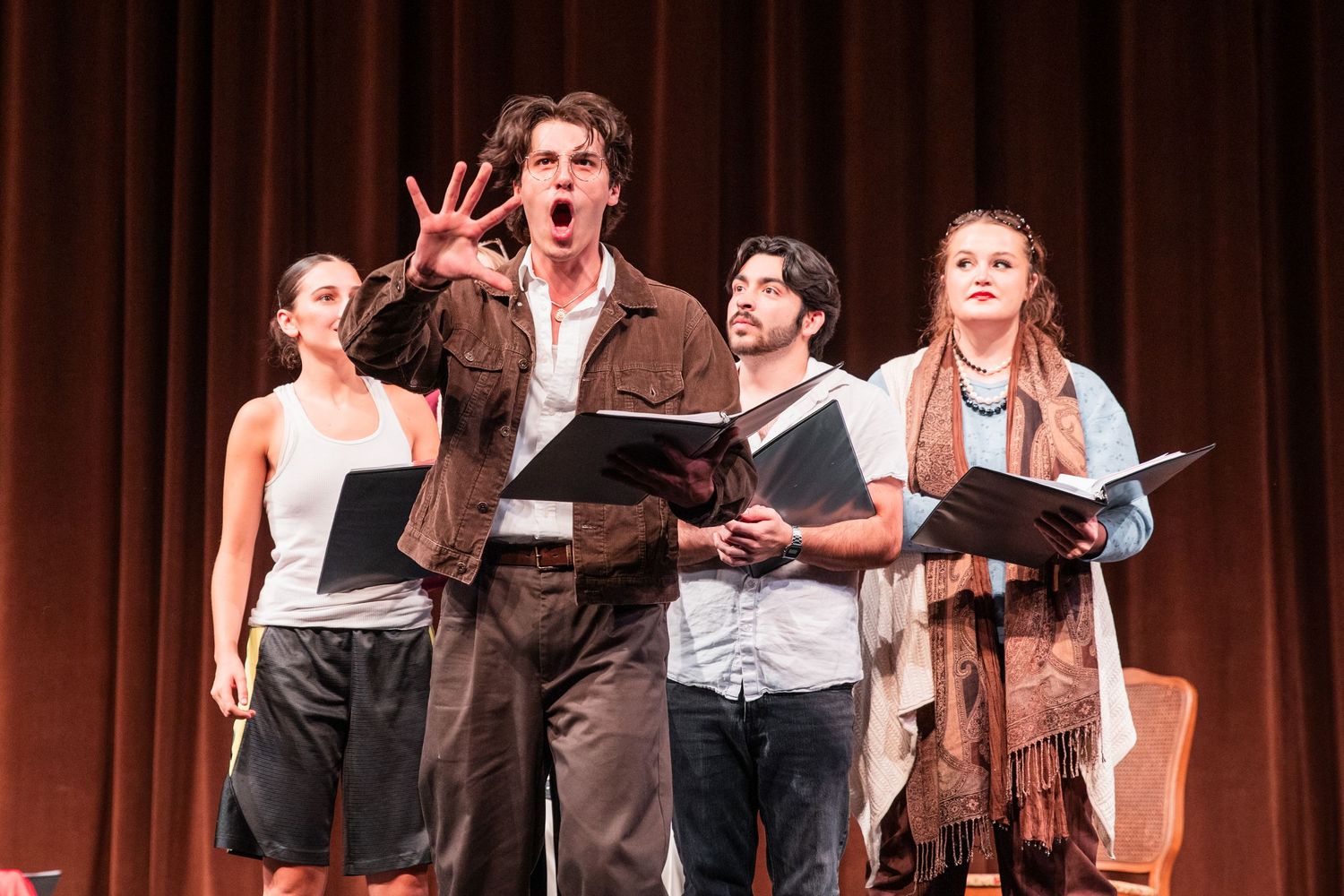 Four theater performers singing expressively on stage holding scripts during a musical production.