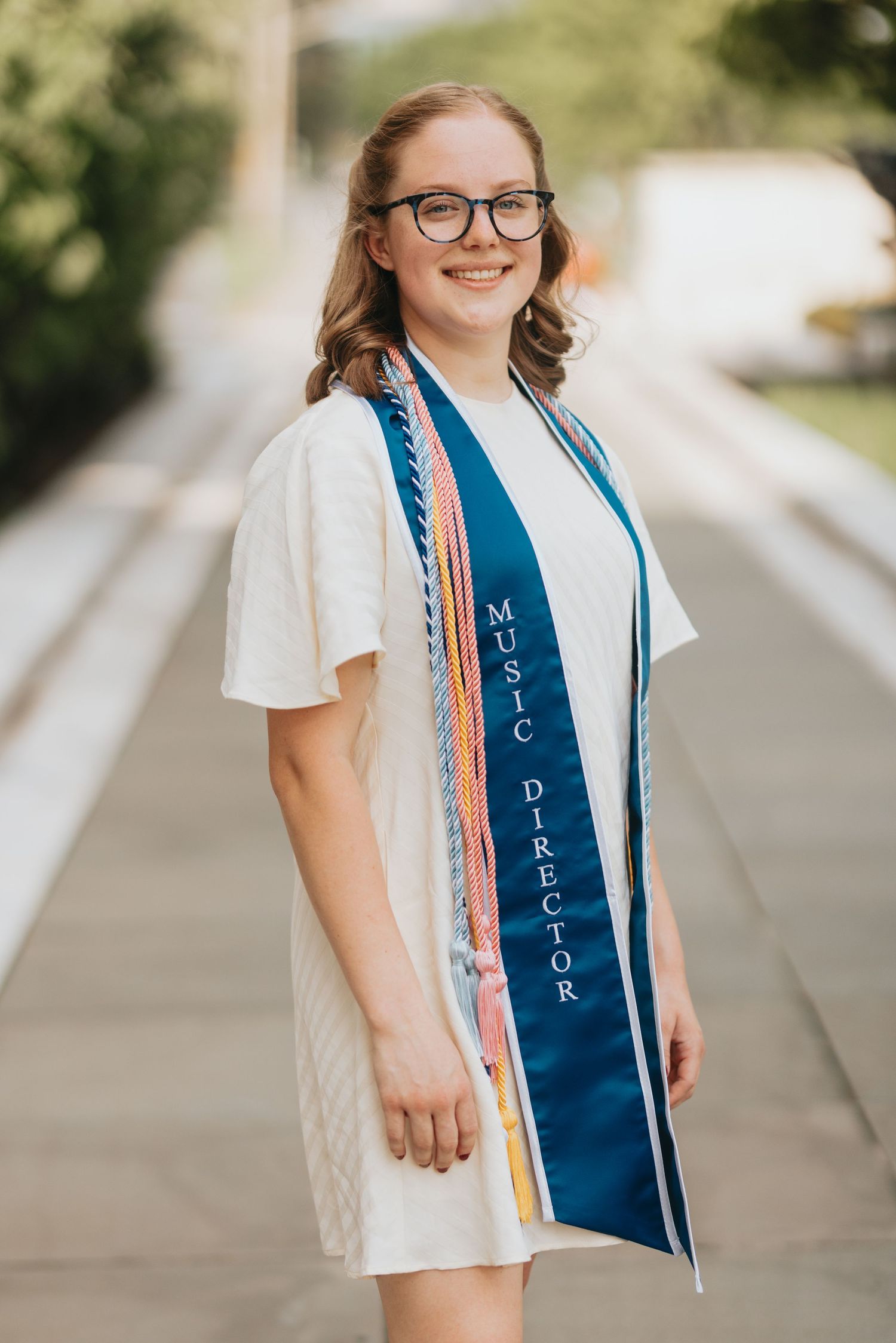 Smiling young woman in white dress wearing Music Director graduation sash outdoors.