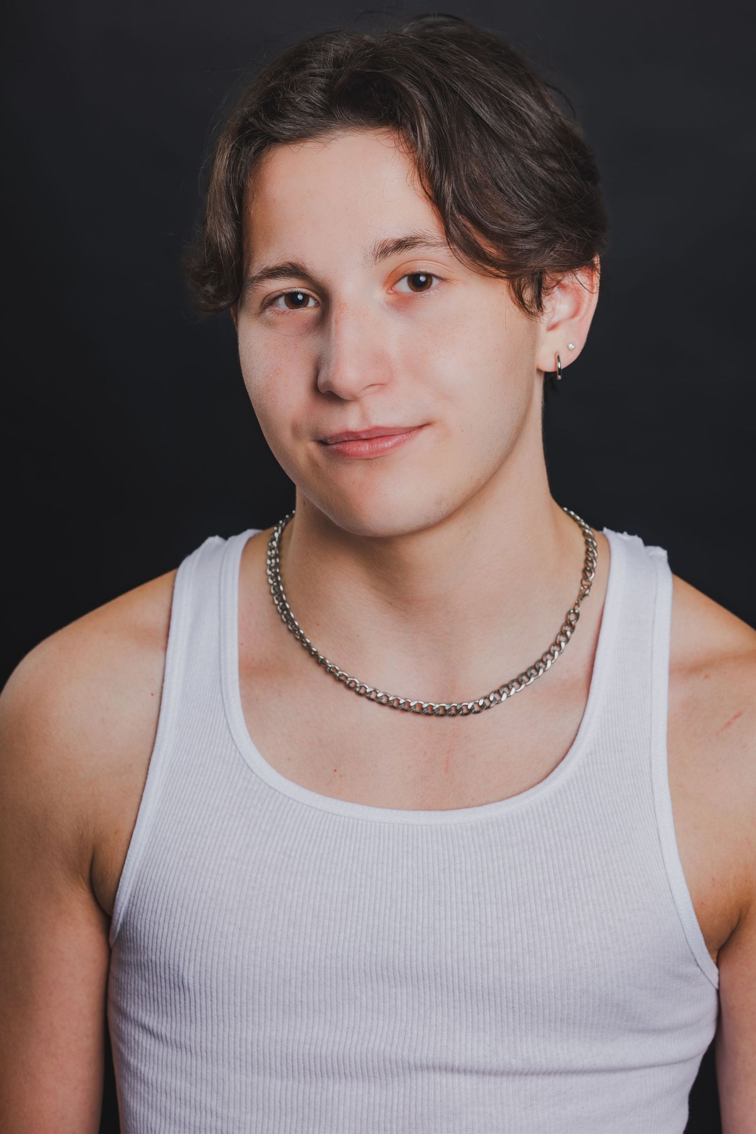 Young person in white tank top with chain necklace against dark background.
