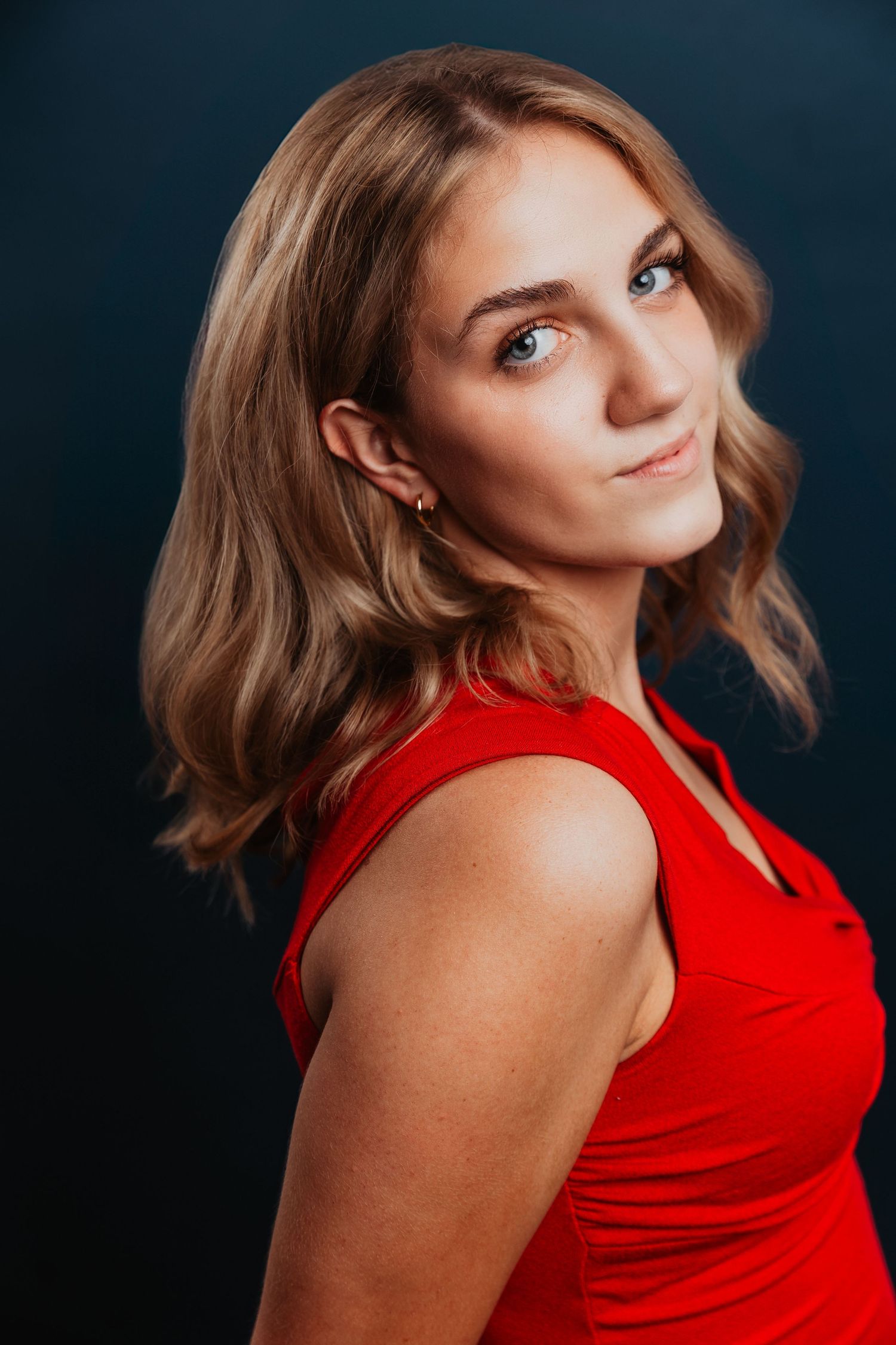 Young woman with wavy blonde hair poses in a red sleeveless dress against a dark background.