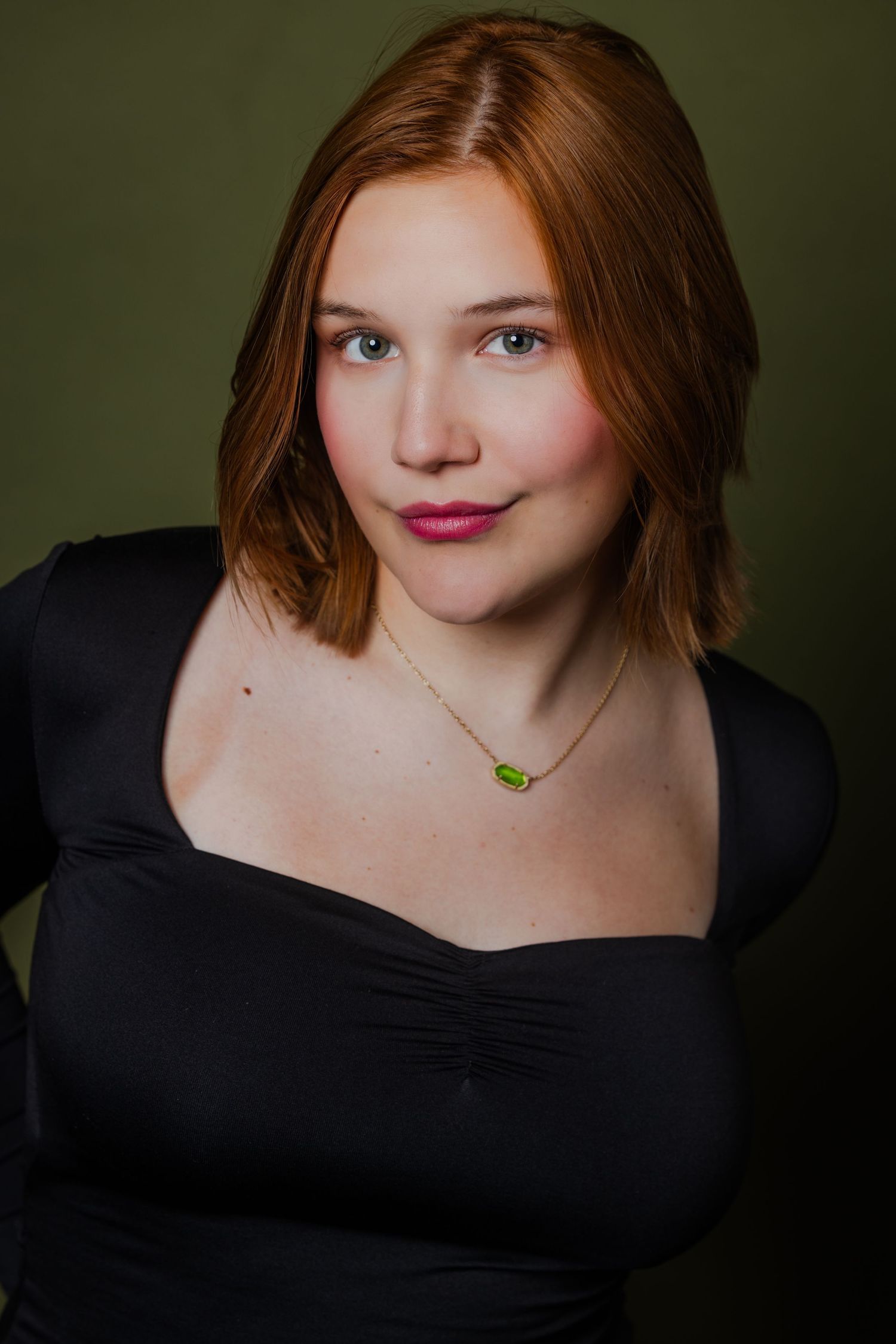 Young woman with red hair wearing a black top and green pendant necklace on a green background.