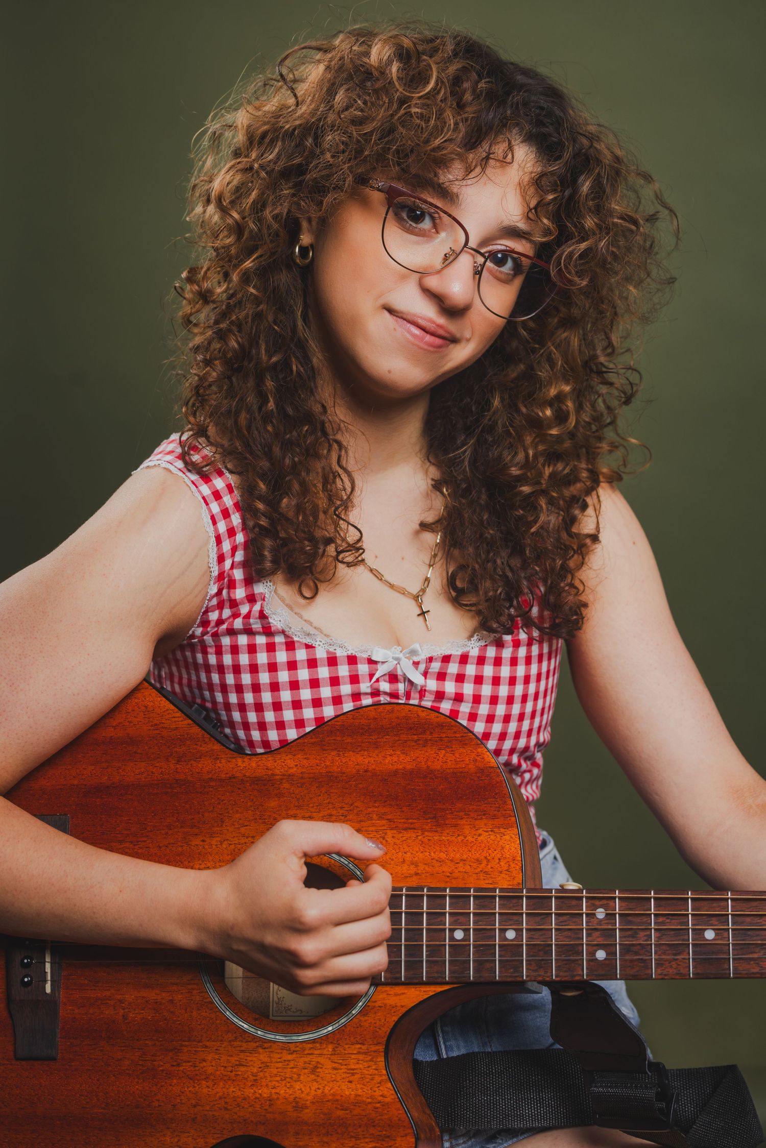 Young woman with curly hair and glasses playing acoustic guitar, wearing a red plaid top in a studio portrait.